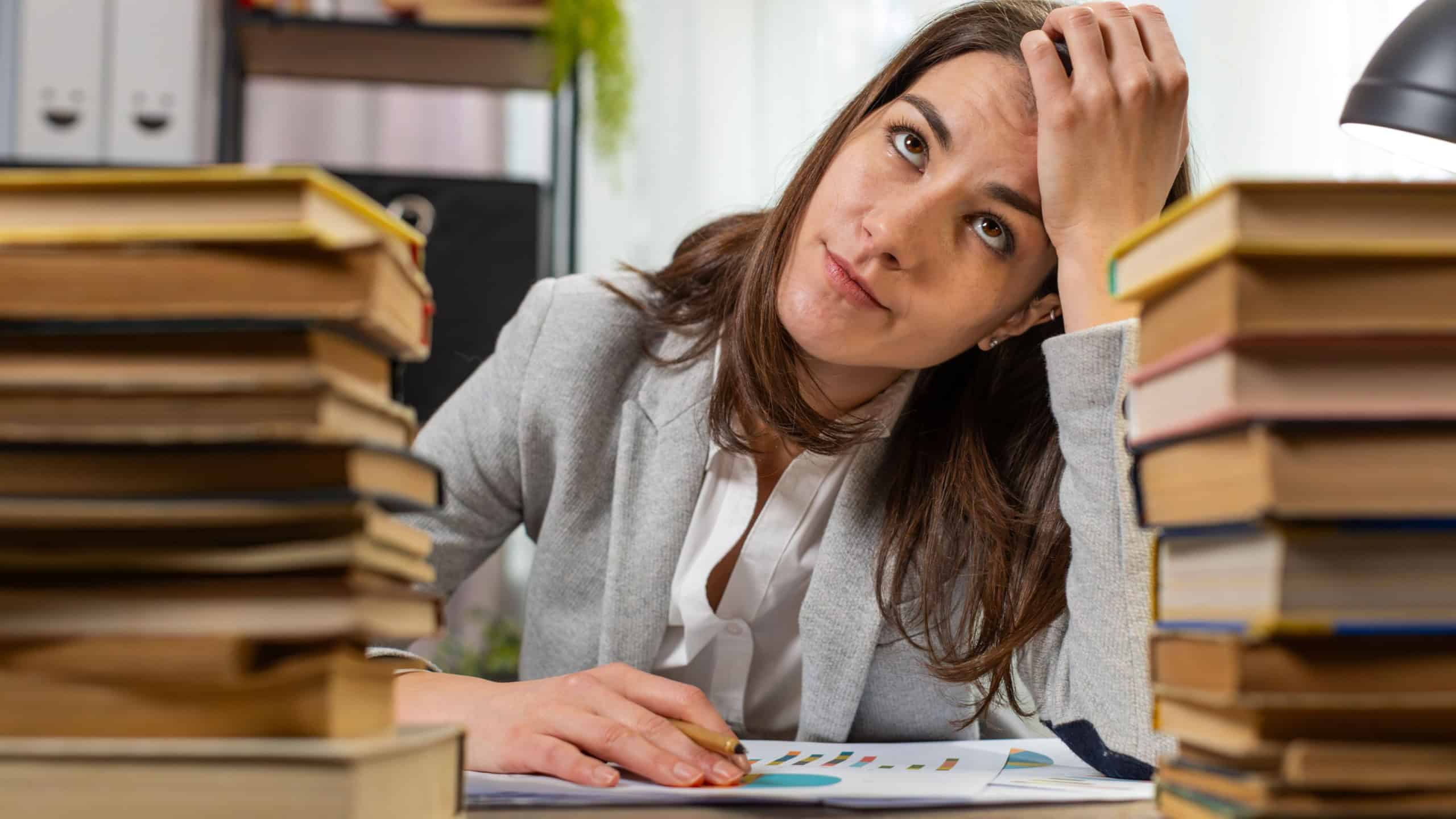 Exhausted Caucasian businesswoman looking at documents reports while working hard at home office desk. Stressed alone girl freelancer touching head in despair surrounded by stacks of books on desk
