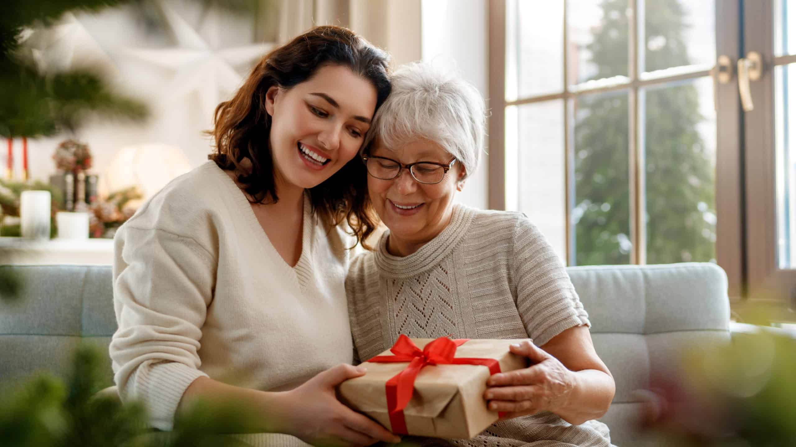 Merry Christmas and Happy Holidays! Senior mom and her adult daughter exchanging gifts. Having fun near tree indoors. 