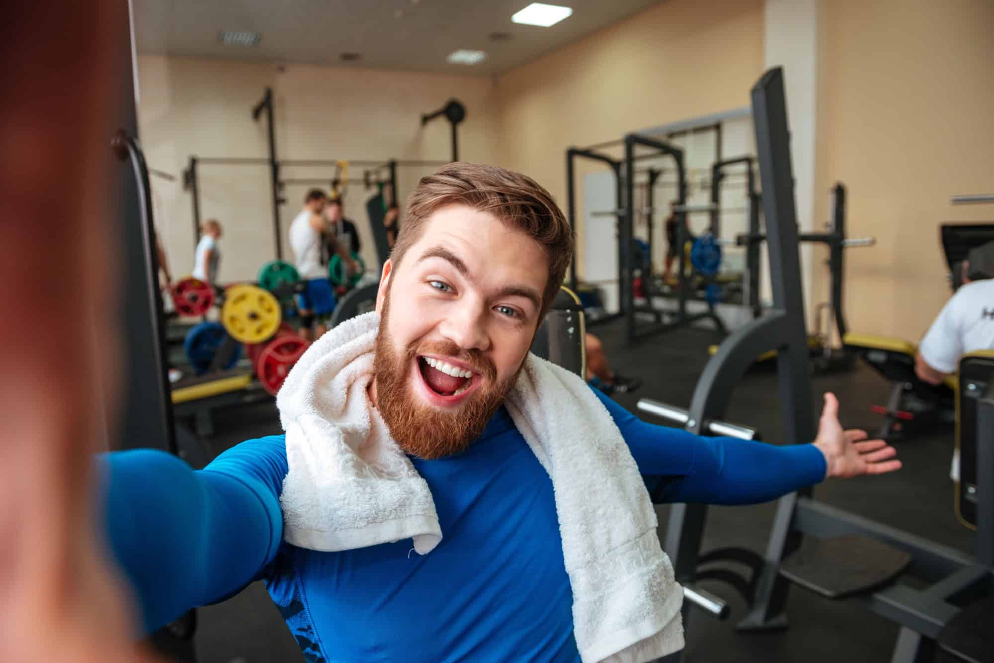 Image of cheerful young sportsman with towel sitting in gym and make selfie.