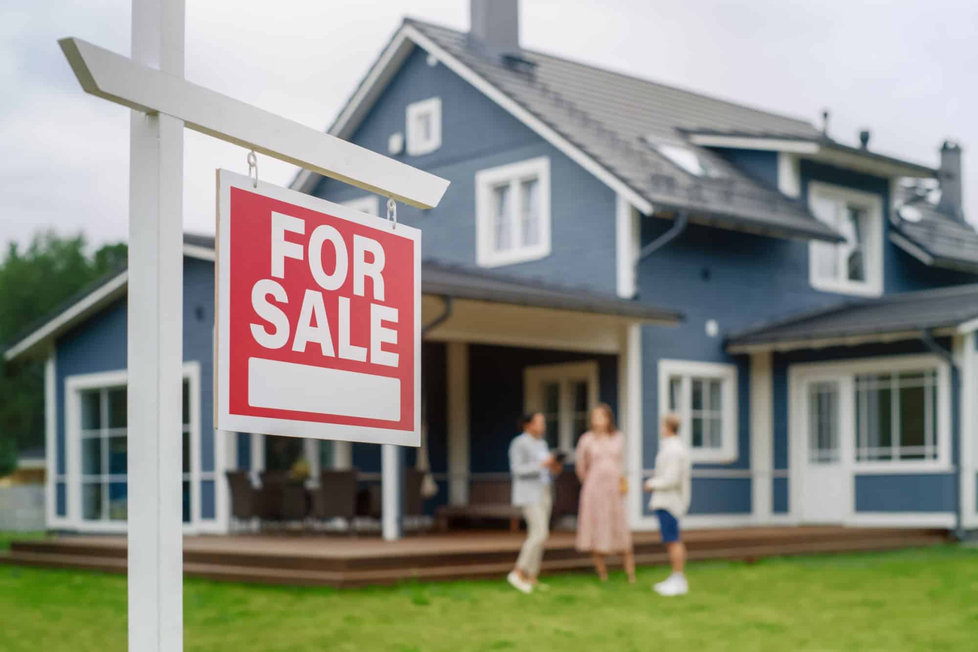 Young Couple Visiting a Potential New Home Property with Professional Real Estate Agent. Female Realtor Showing the Area to Future Homeowners. Focus on For Sale Sign.