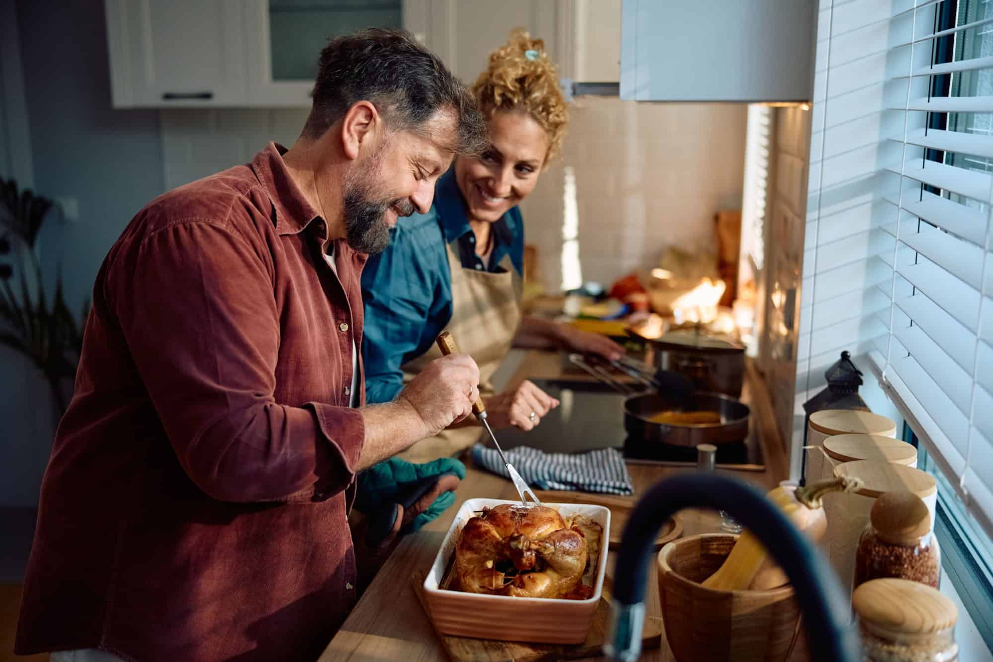 Happy man and his wife preparing roast turkey for Thanksgiving meal while cooking together in the kitchen.