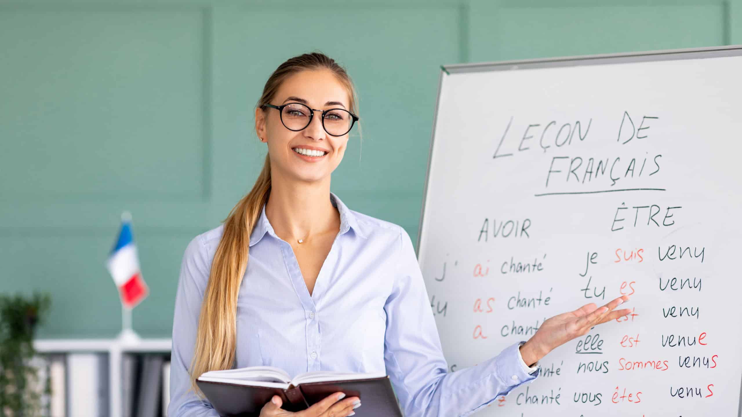Happy French teacher explaining foreign language rules near blackboard indoors and smiling at camera. Confident young female tutor teaching linguistic online course