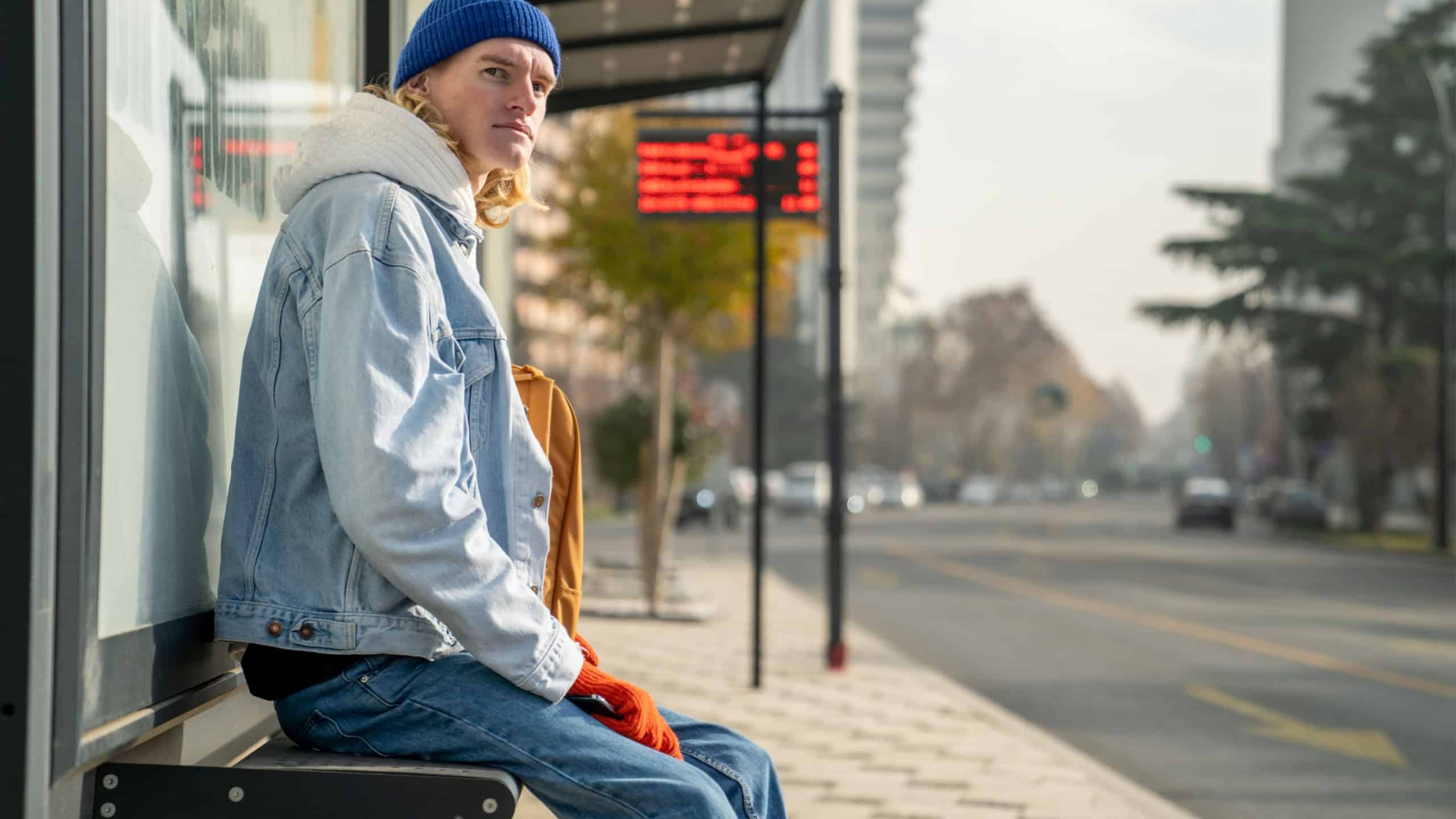 Blond guy student waiting transport very long time in morning. Man on bus stop in sunny weather. Blonde hair millennial man on bus stop in city. Modern urban transport for everyone