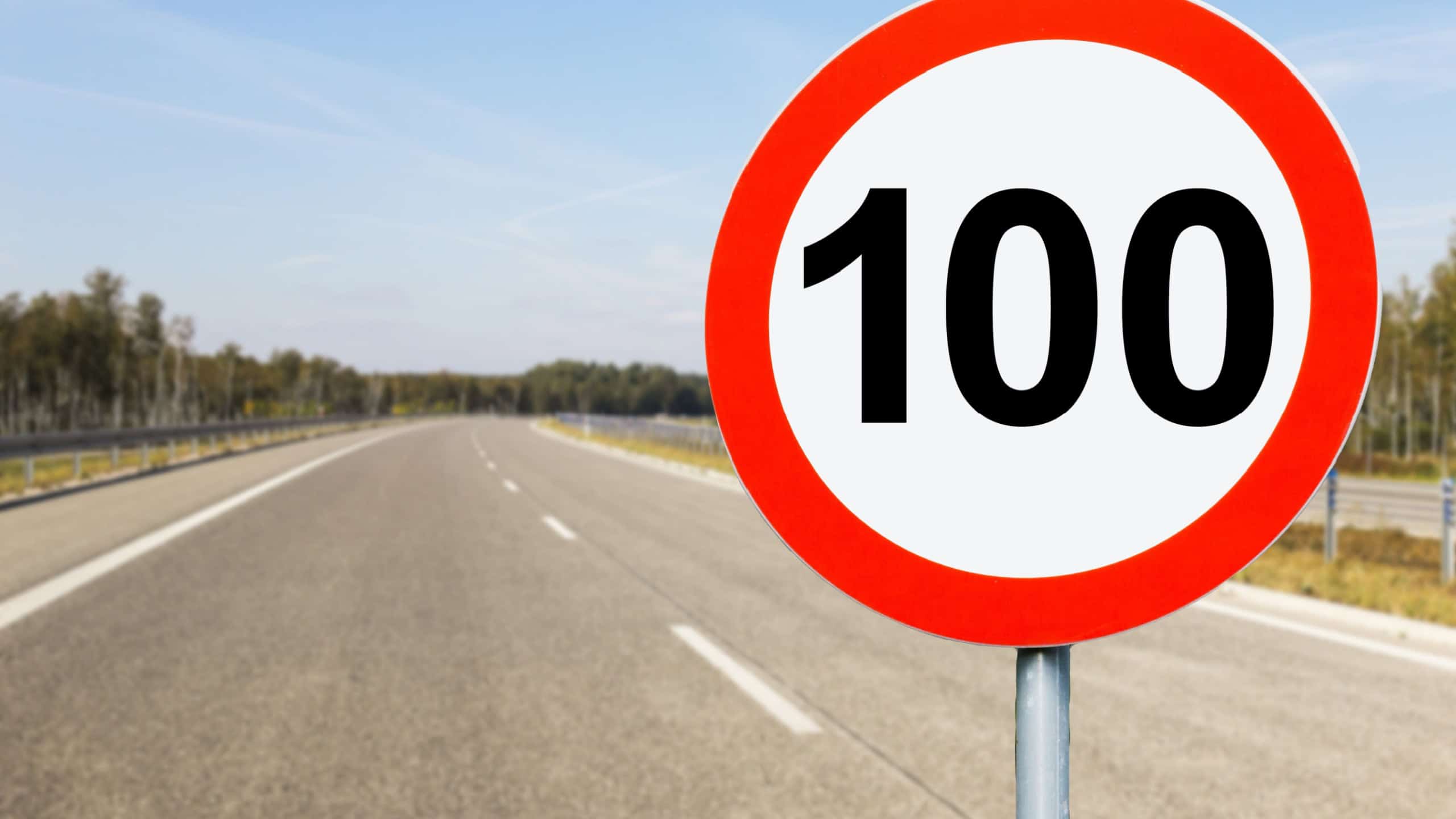 Speed limit sign background. 100 kmph on a highway. Multiband highway empty road. Driving under a bright blue sky. Overpass and roadside view on a clear day in Europe. Asphalt road landscape.