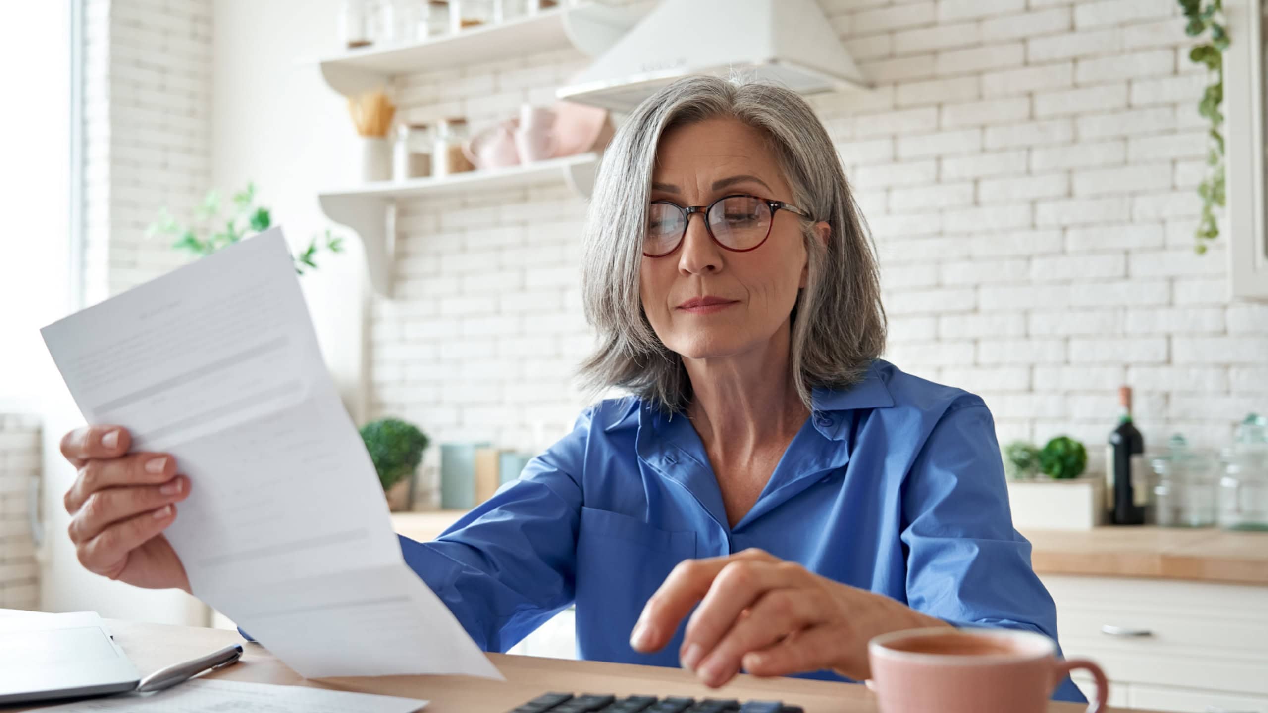 Senior mature business woman holding paper bill using calculator, old lady managing account finance, calculating money budget tax, planning banking loan debt pension payment sit at home kitchen table.