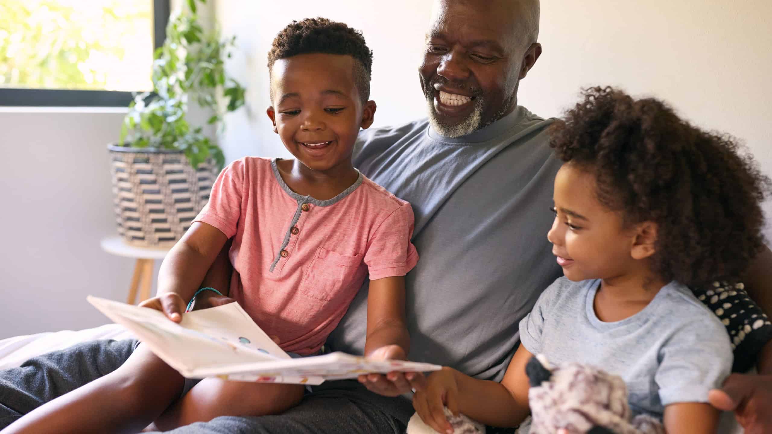 Grandfather Sitting On Sofa At Home Reading Book With Grandchildren