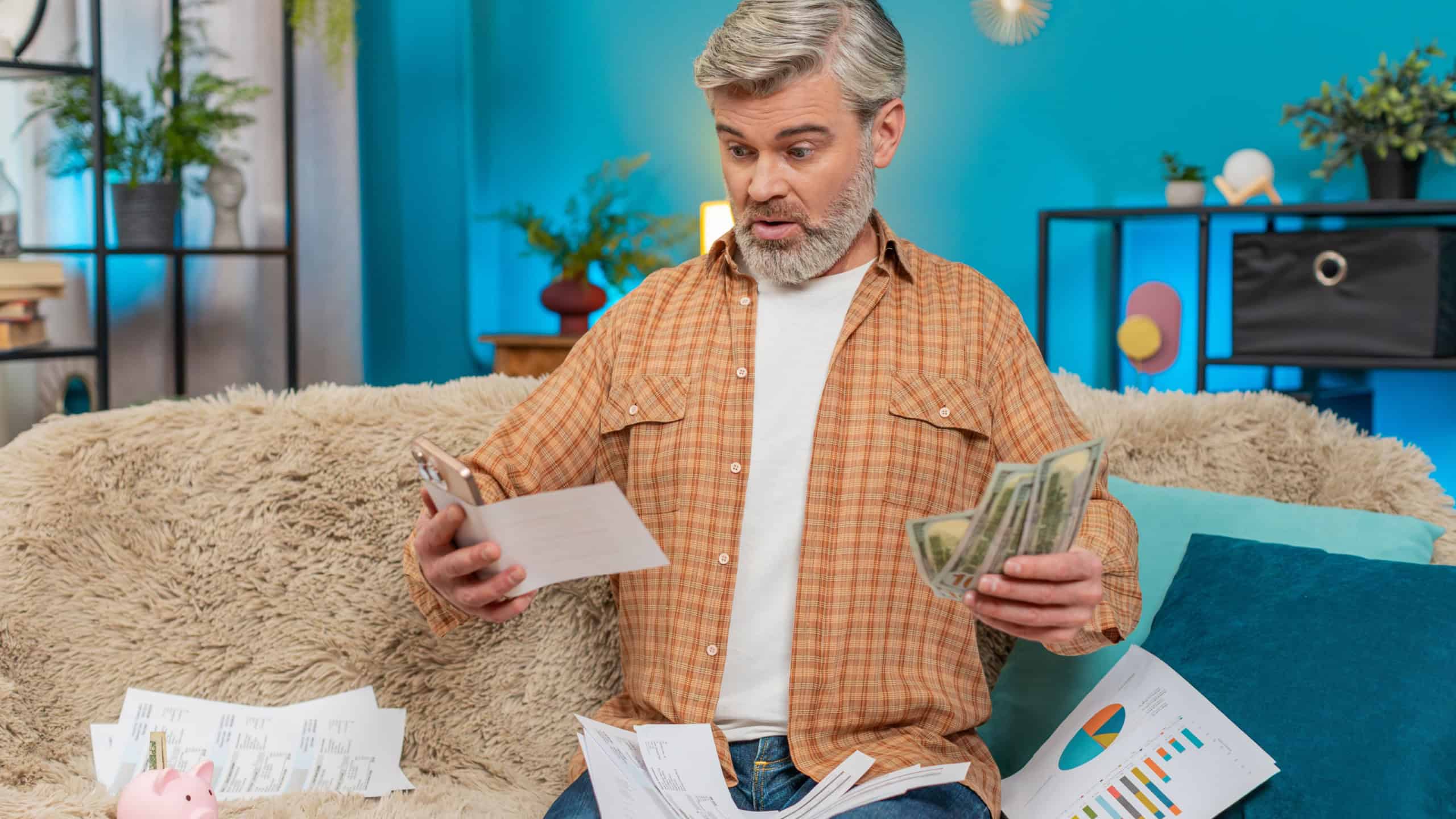 Mature man sitting on home sofa, using smartphone with calculator app to count money dollars cash. Guy surrounded by documents, invoices, bills and a piggy bank, exhausted from financial calculations