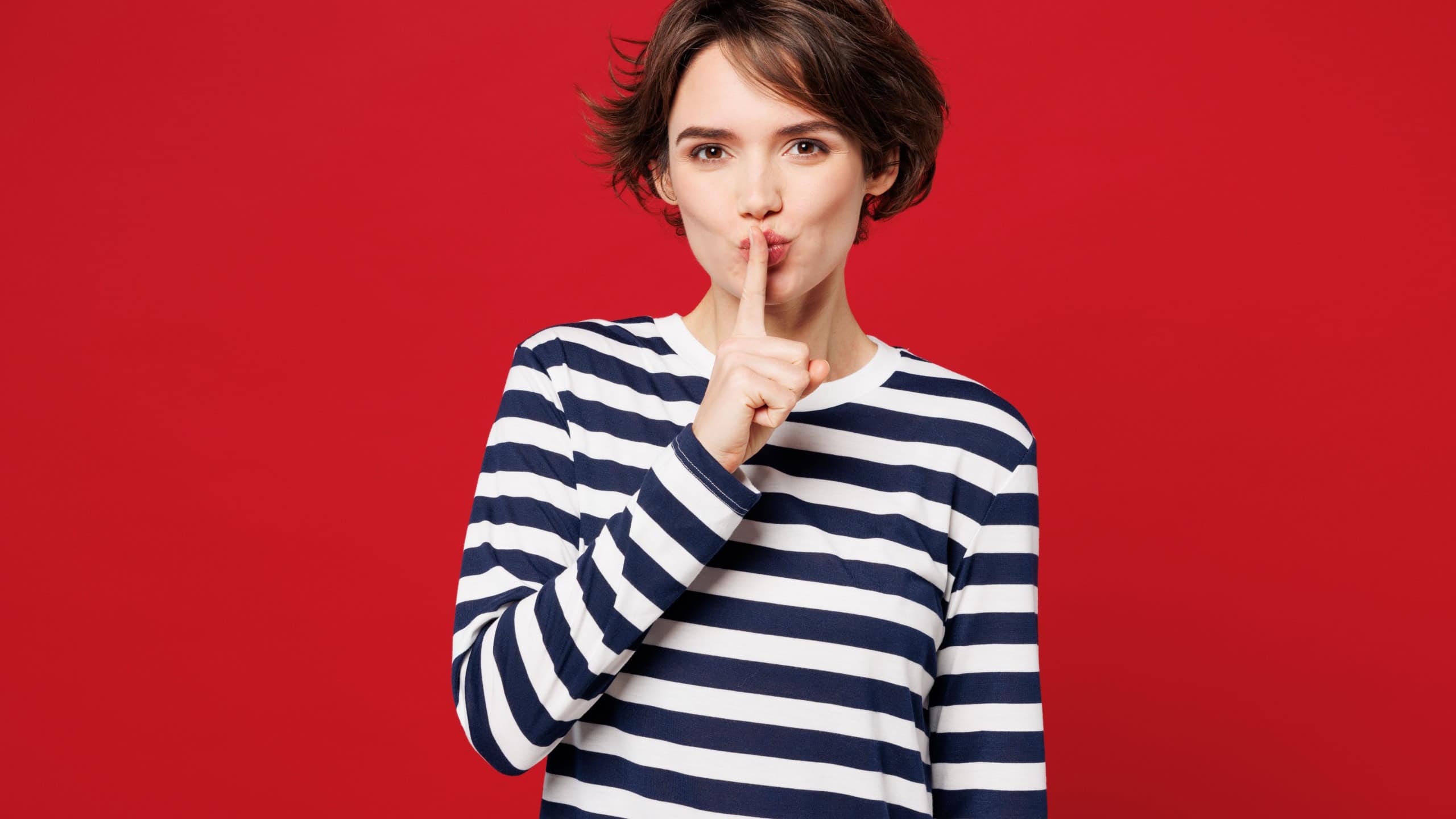 Young secret happy woman wear blue striped sweatshirt casual clothes say hush be quiet with finger on lips shhh gesture look camera isolated on plain red background studio portrait. Lifestyle concept