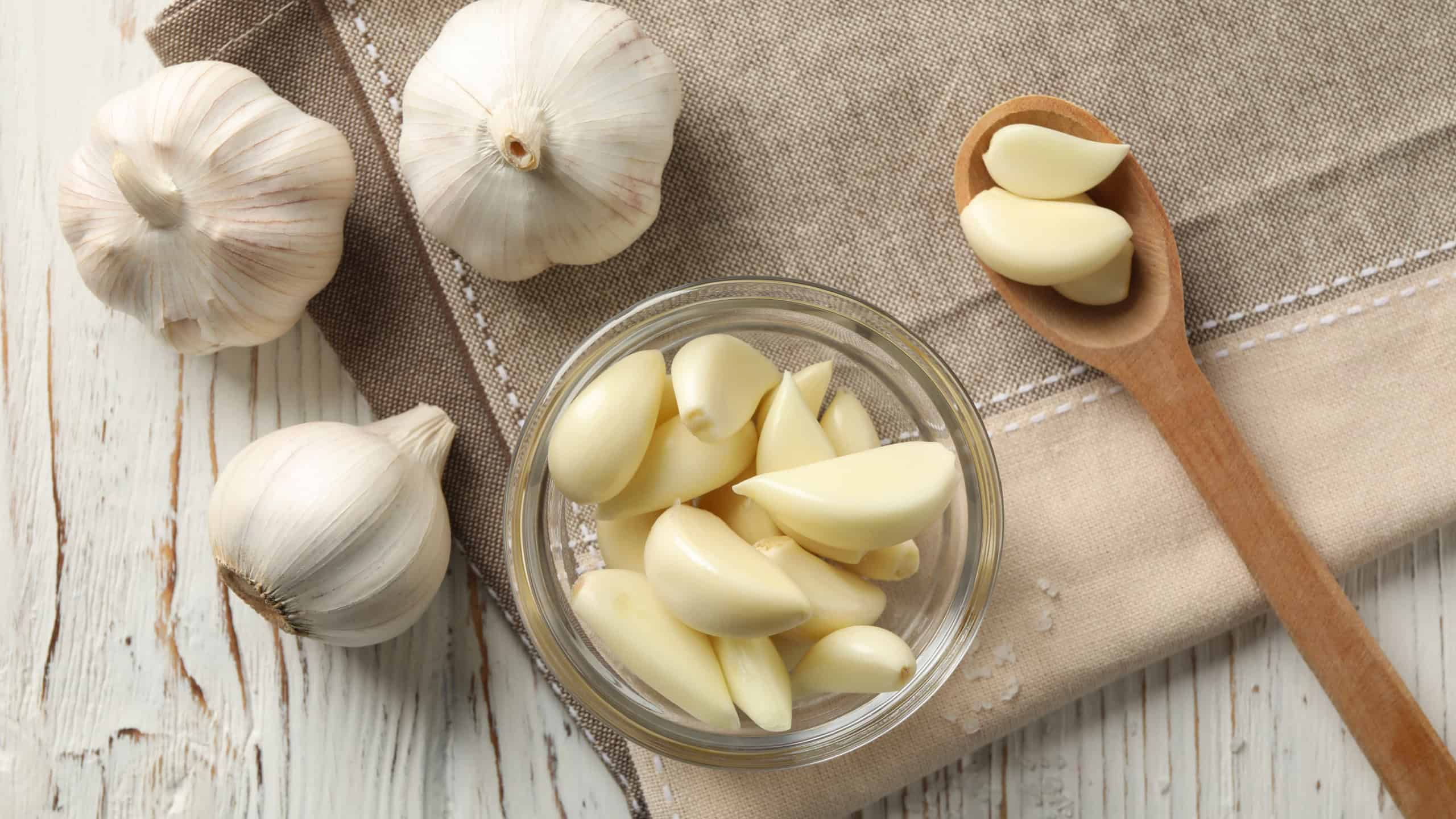 Glass bowl of fresh garlic bulbs, spoon of slices, napkin on white wooden background, closeup. Top view