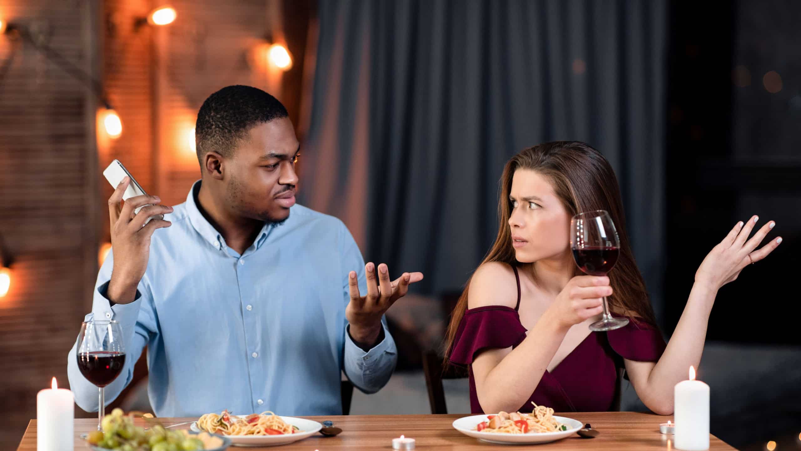Young Woman Complaining About Her Black Boyfriend Talking On Cellphone During Romantic Date In Restaurant, Feeling Annoyed And Bored. Interracial Couple Having Problems In Relationship, Free Space
