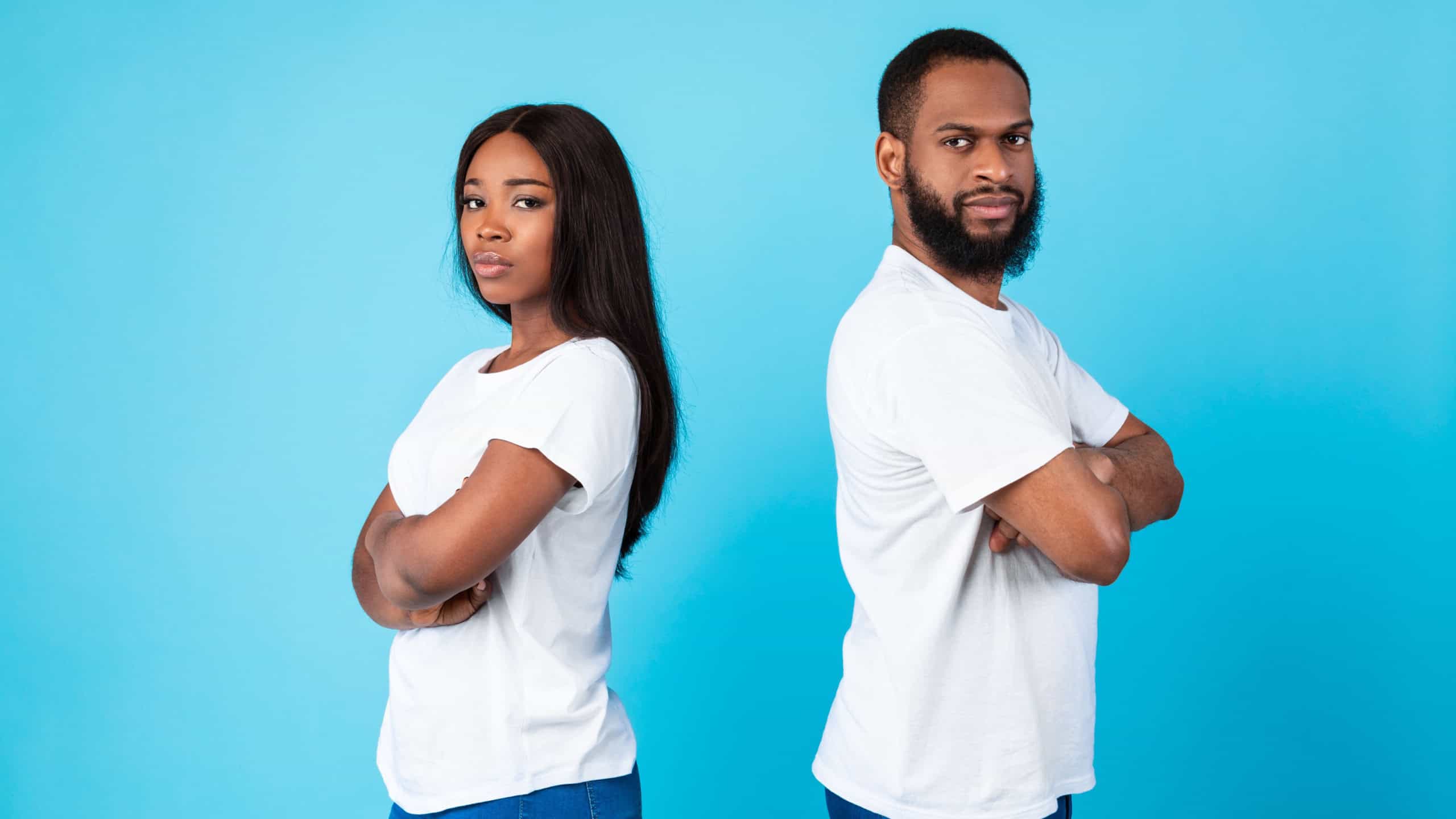 Portrait of serious African American millennial couple standing separate back to back with folded arms, black guy and lady looking at camera isolated over blue studio background wall