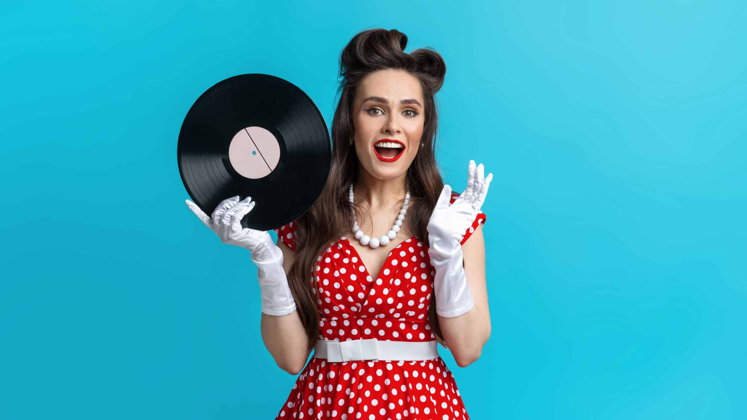 Retro music, old songs, nostalgia. Excited young pinup woman in retro polka dot dress holding gramophone vinyl record, smiling at camera over blue studio background