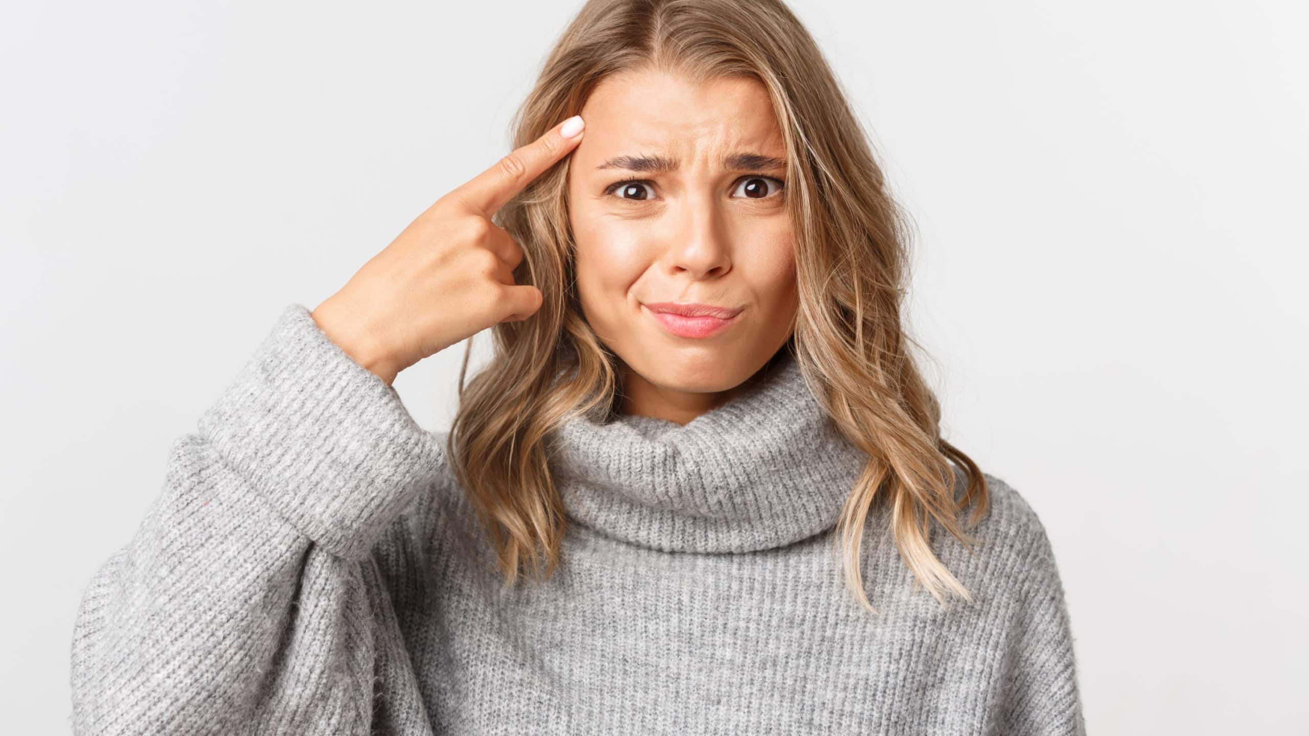 Close-up of confused blond girl in grey sweater, pointing finger at head, scolding something for acting stupid, standing over white background