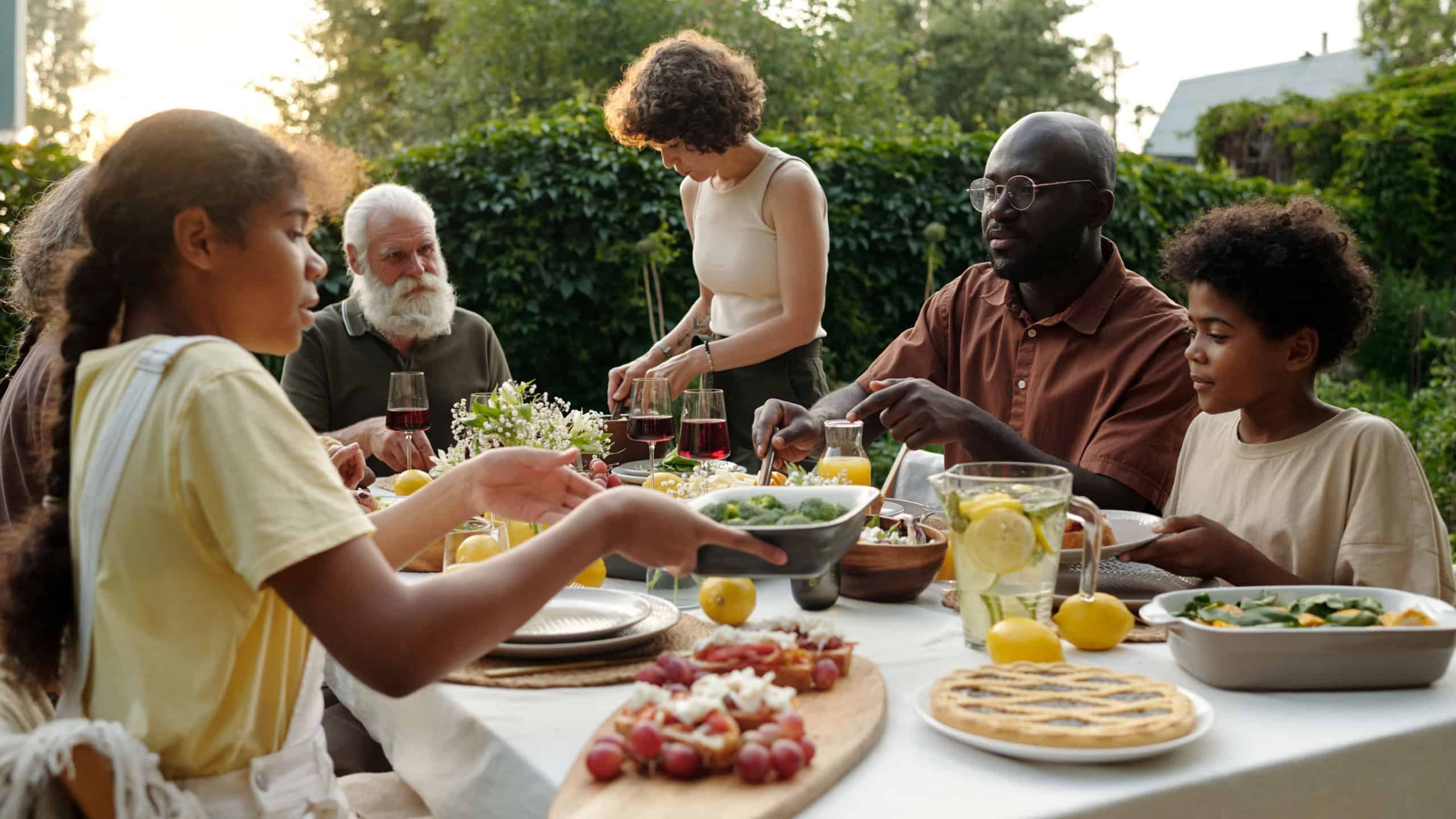 Modern family of siblings, their intercultural parents and grandparents sitting by dinner table served with homemade food and drinks