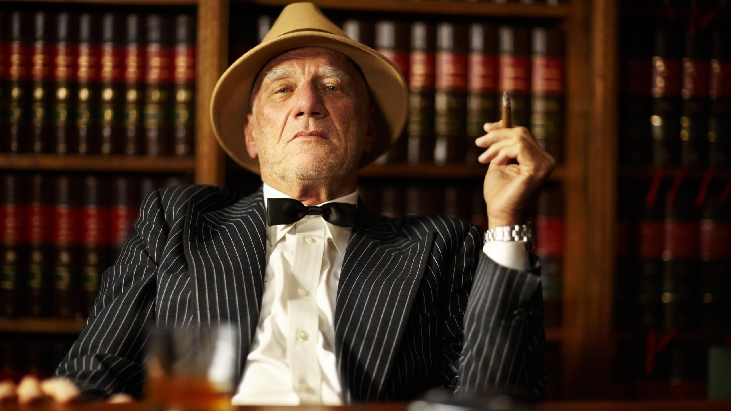 Portrait, mafia and boss with cigarette in office for habit, smoking or relaxing by desk. Mature, mob leader and retro fashion in suit, bowtie and hat for crime, danger or power with vintage look