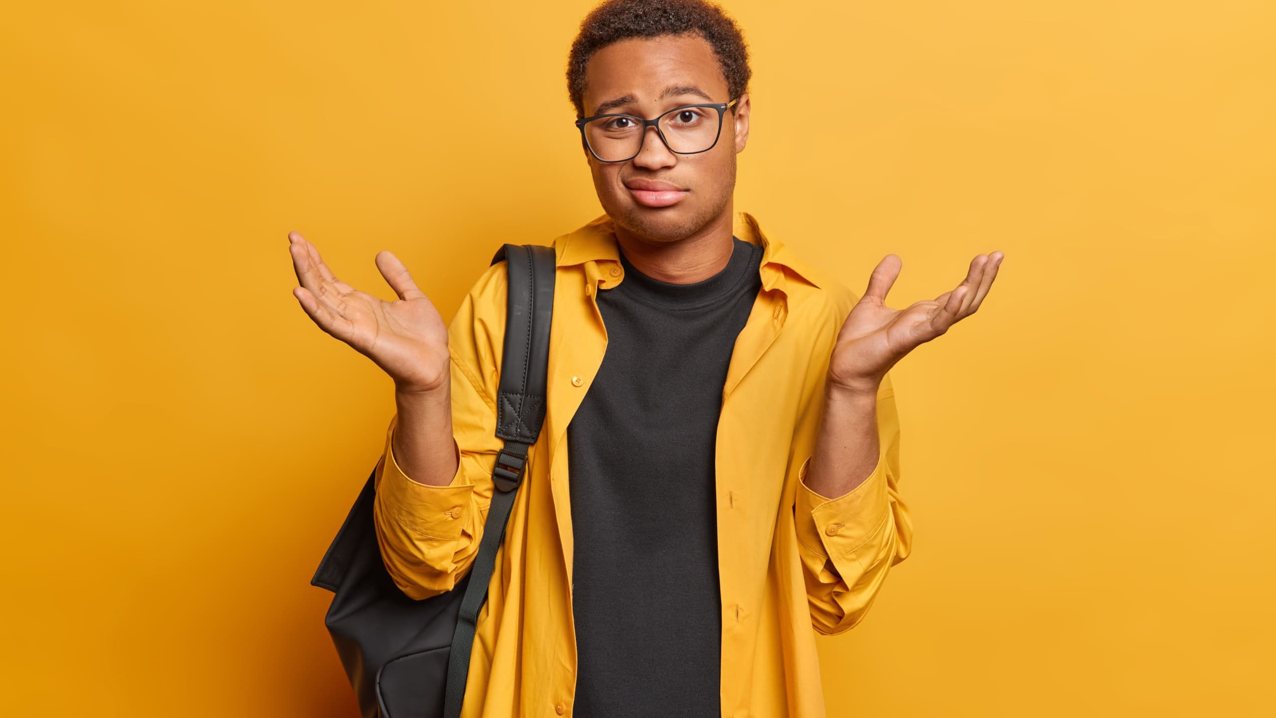 People emotions concept. Indoor shot of young confused African american male wearing casual clothes posing in centre isolated on yellow background spreading palms with hesiatation shrugging shoulders
