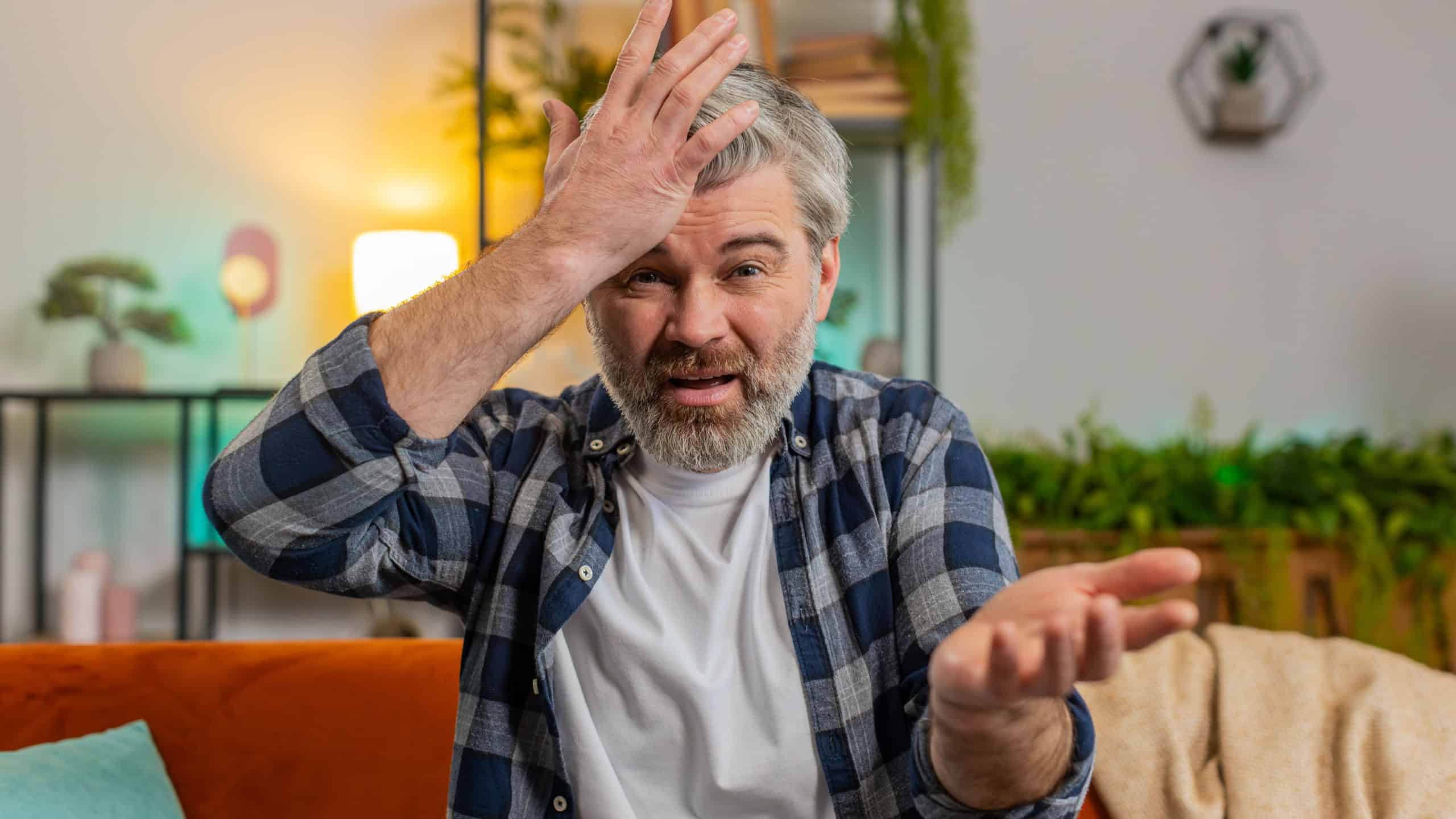 Dissatisfied middle-aged gray haired bearded man arguing quarrel talking on video call sitting on sofa in room at home. Angry mature male guy complains about problems looking in camera