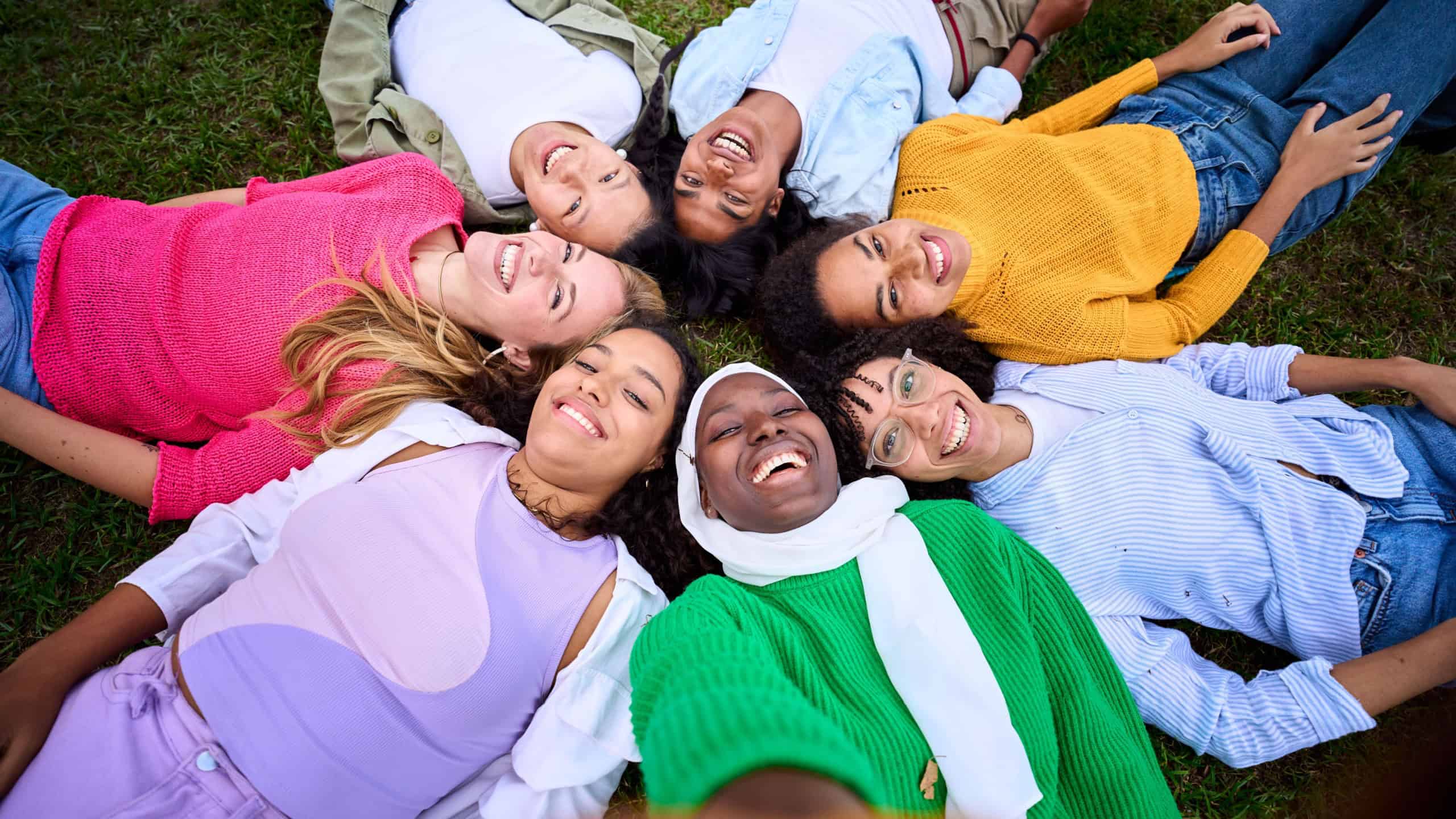 Selfie of multiracial group young female community friends lying in circle on grass enjoying together looking smiling at camera. Diverse happy only women posing joyful bonding together at city park
