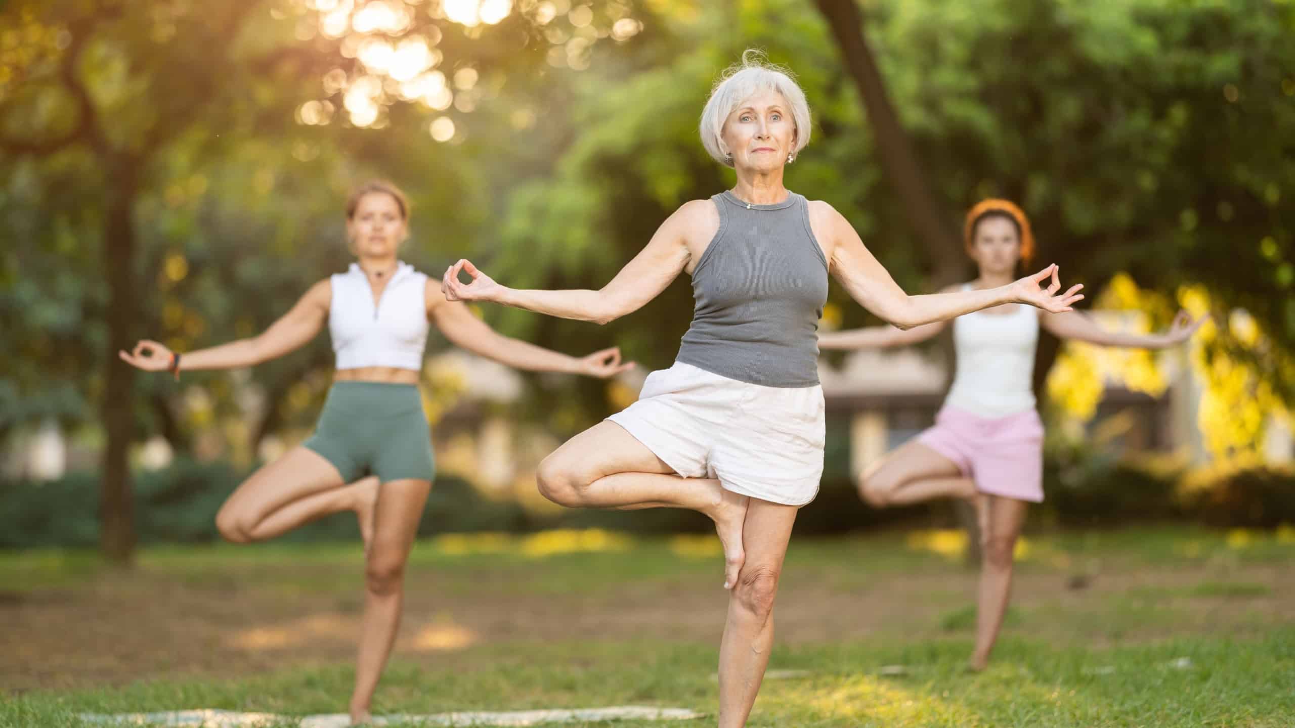 Positive fit senior yogini practicing hatha yoga on shaded glade in green summer park with group of women, doing Vrikshasana asana balancing on one leg