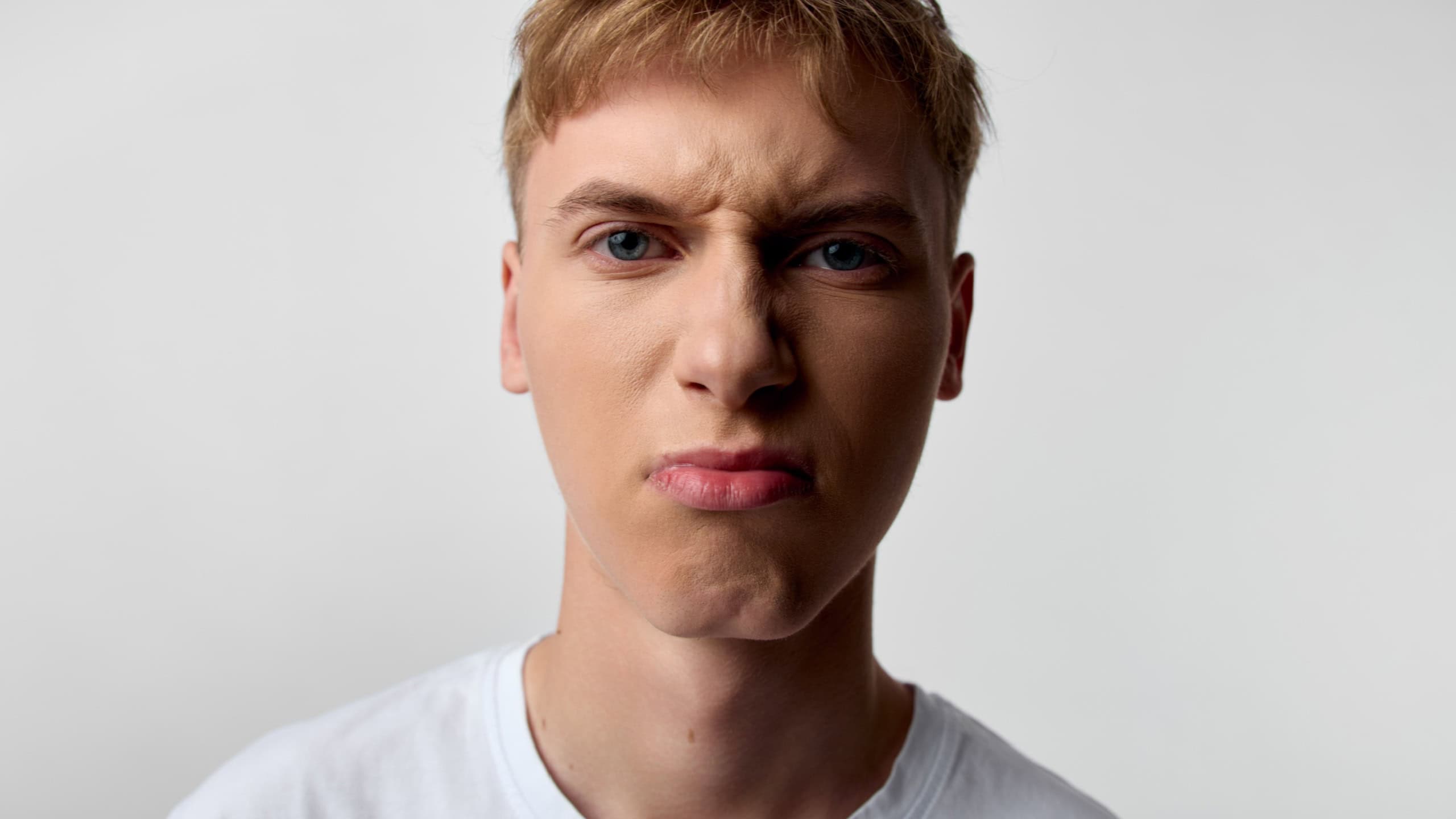 Close up of an annoyed man with angry facial expression, pursed lips, and frustrated gaze, wearing a white t shirt against a plain backdrop, conveying irritation, mood, and intense emotion suitable