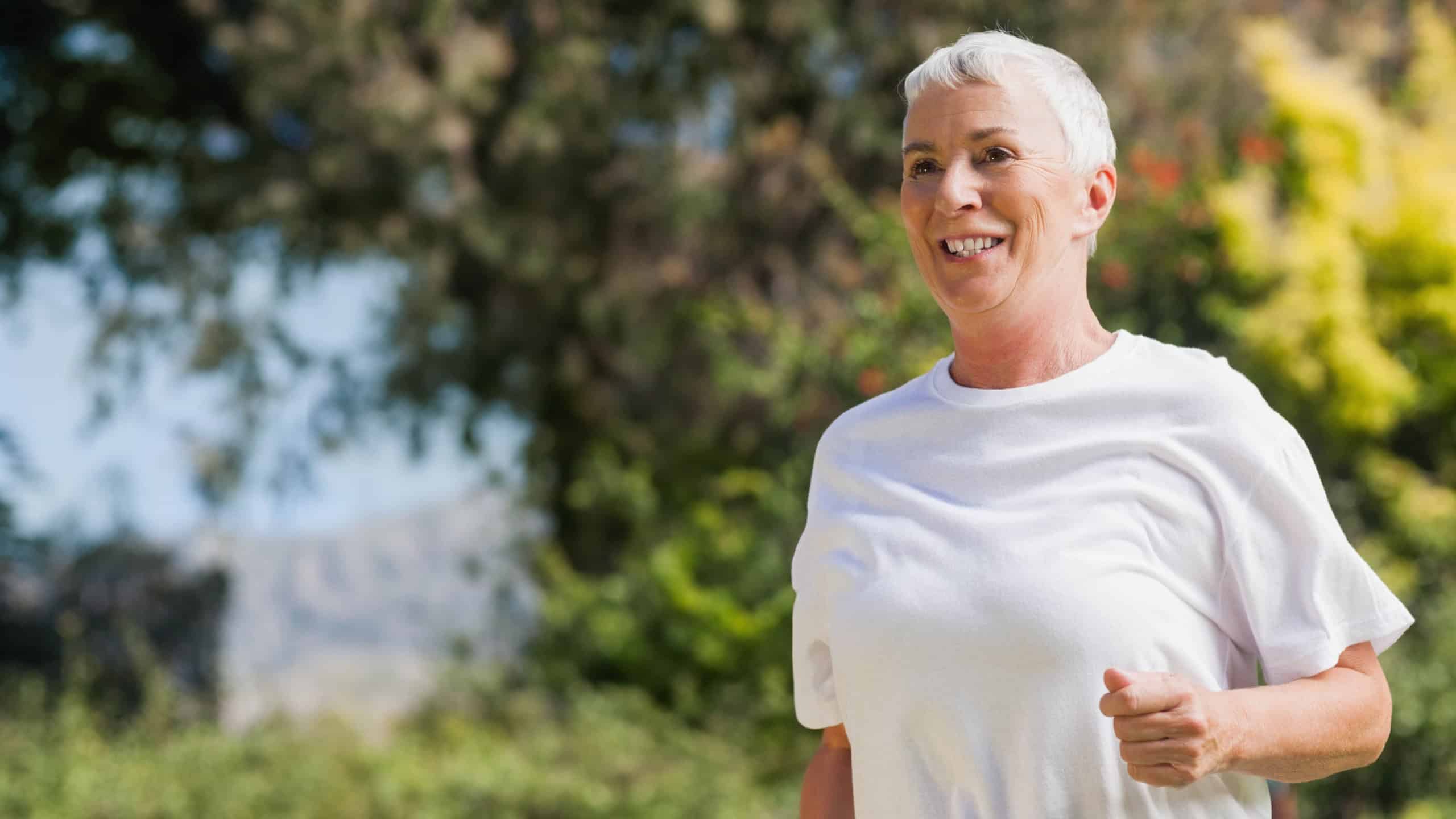 Senior female jogging briskly on park trail wearing white T-shirt with sunlit greenery, copy space. Elderly, outdoors, fitness, photorealistic, advertising, nature, vitality