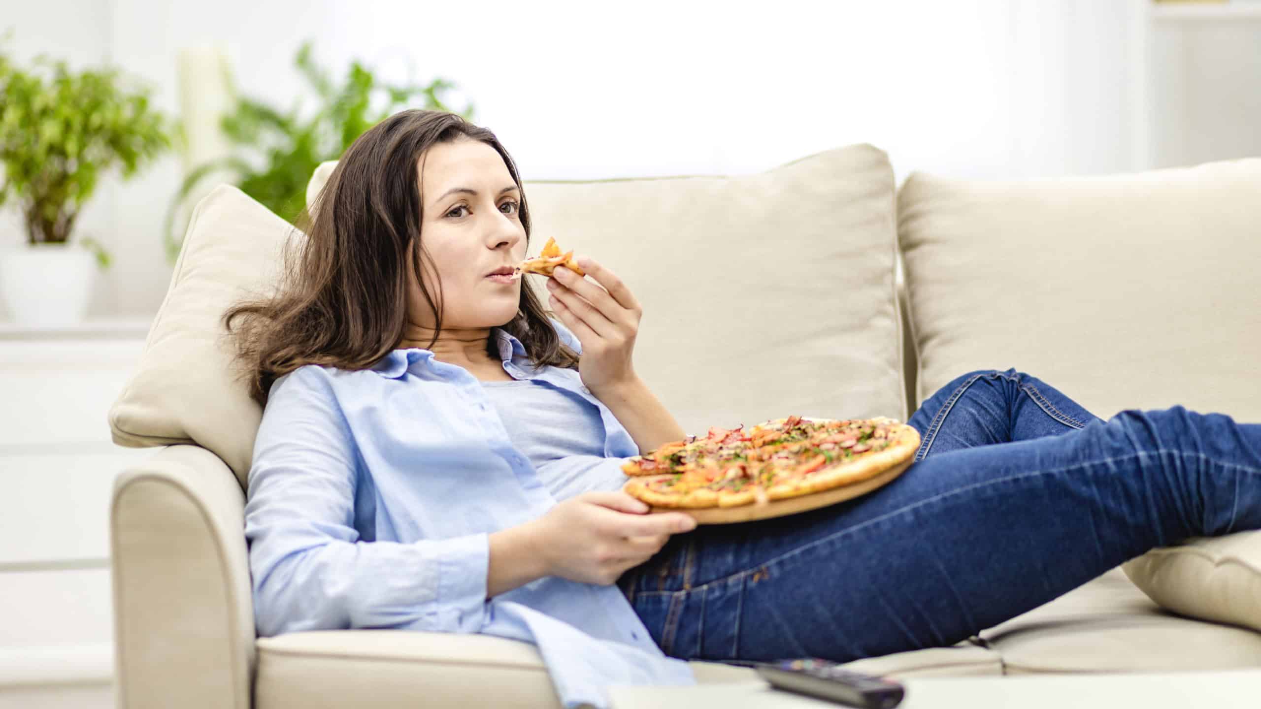 Tired woman is chewing pizza, while laying on the white sofa. She is watching TV shows, being on blurred background.