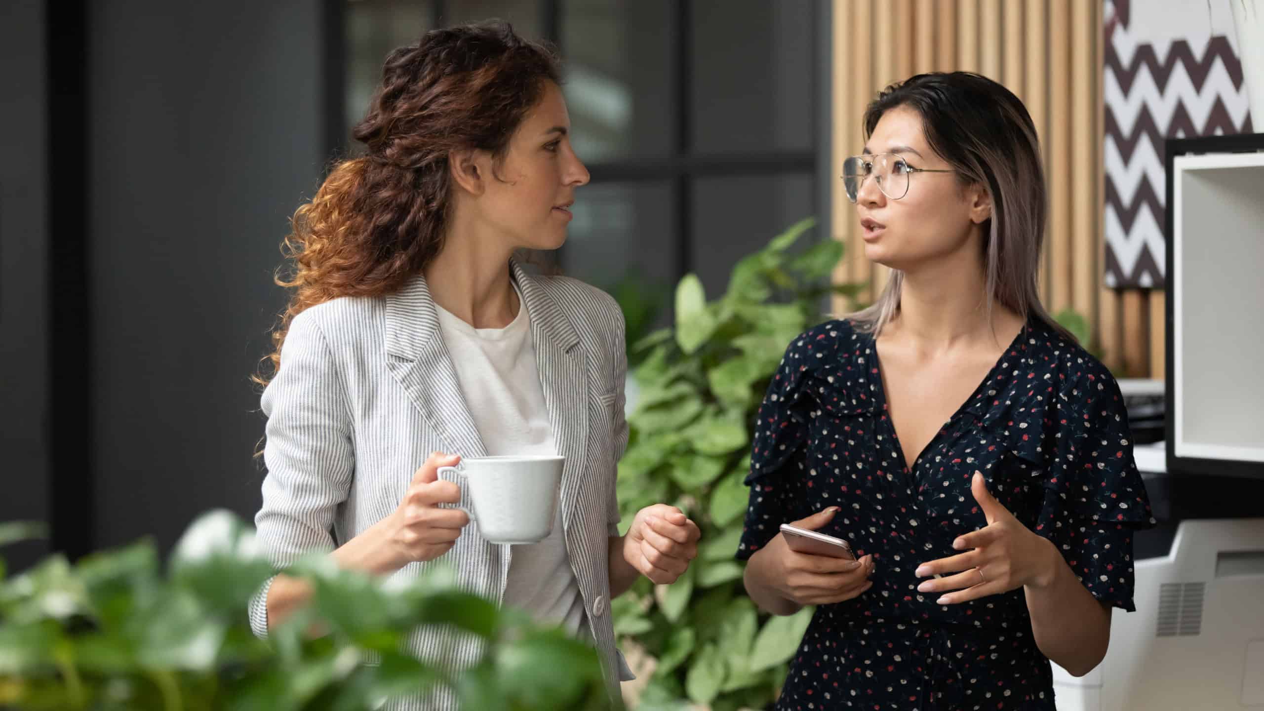 Diverse employees chatting during coffee break, walking in modern office, Asian businesswoman wearing glasses sharing ideas, discussing project with colleague, having pleasant conversation