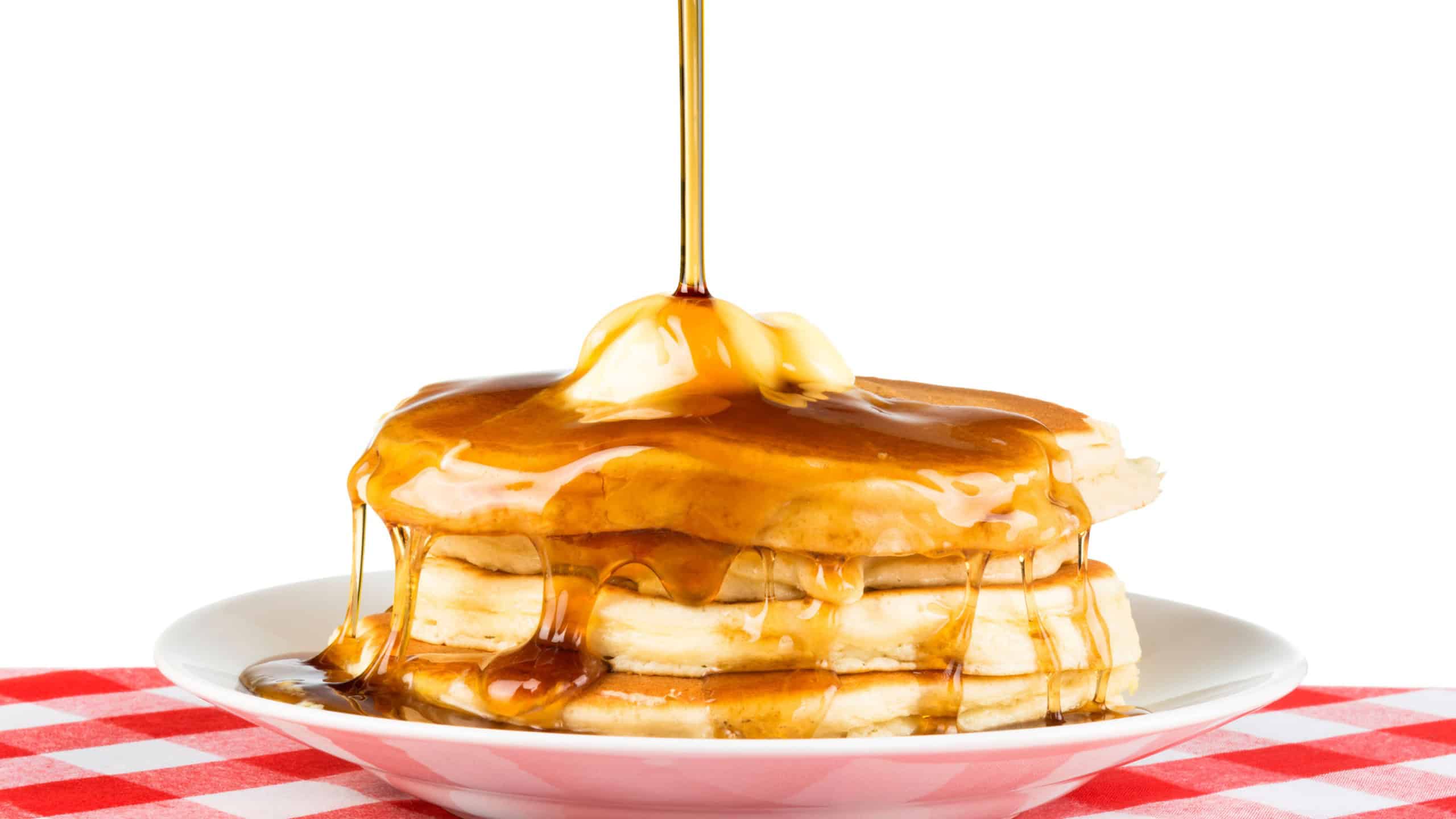 Sweet maple syrup being poured over a hot, freshly buttered stack of pancakes.