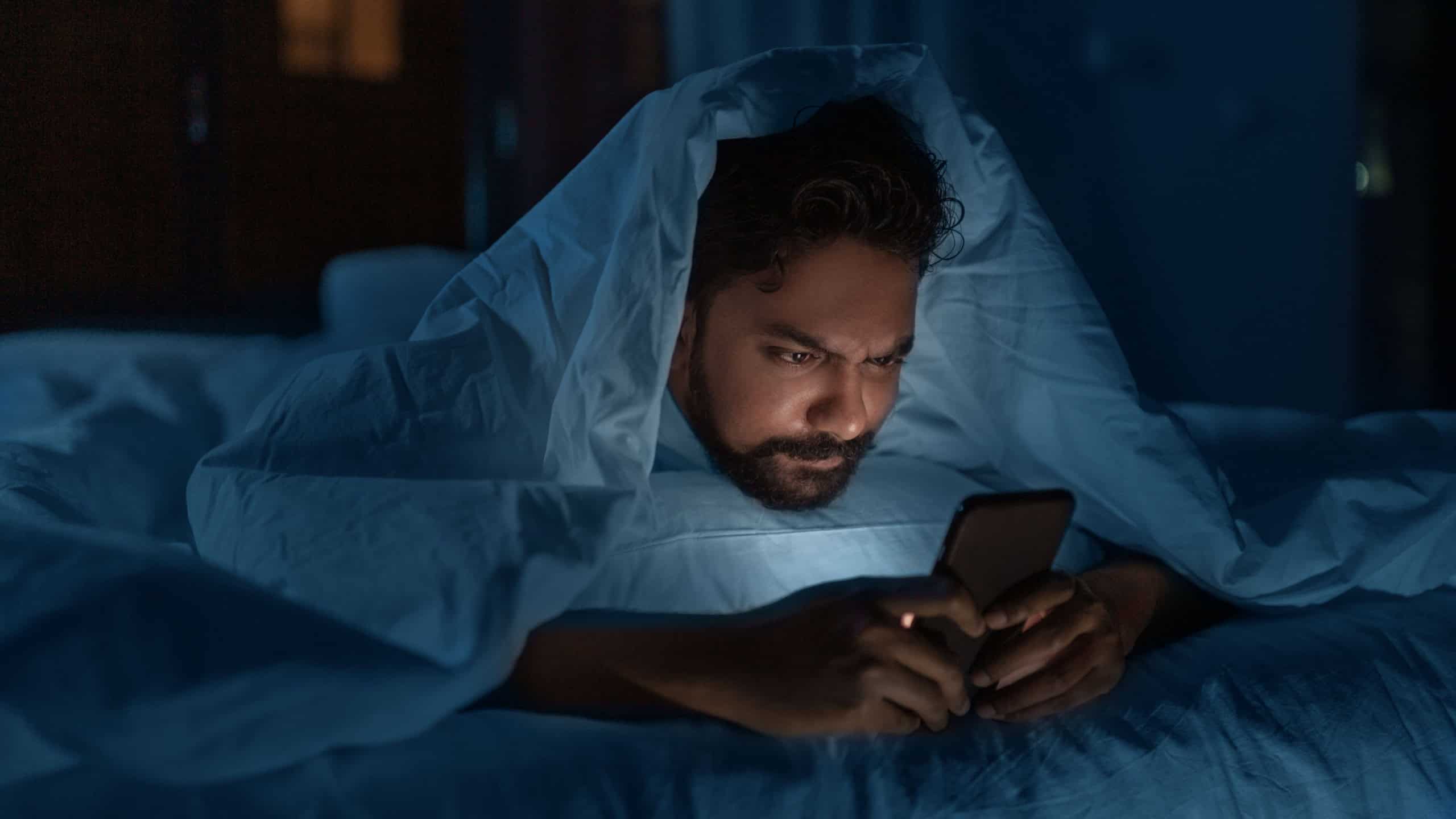 technology, internet, communication and people concept - young indian man with smartphone lying in bed under blanket at home at night