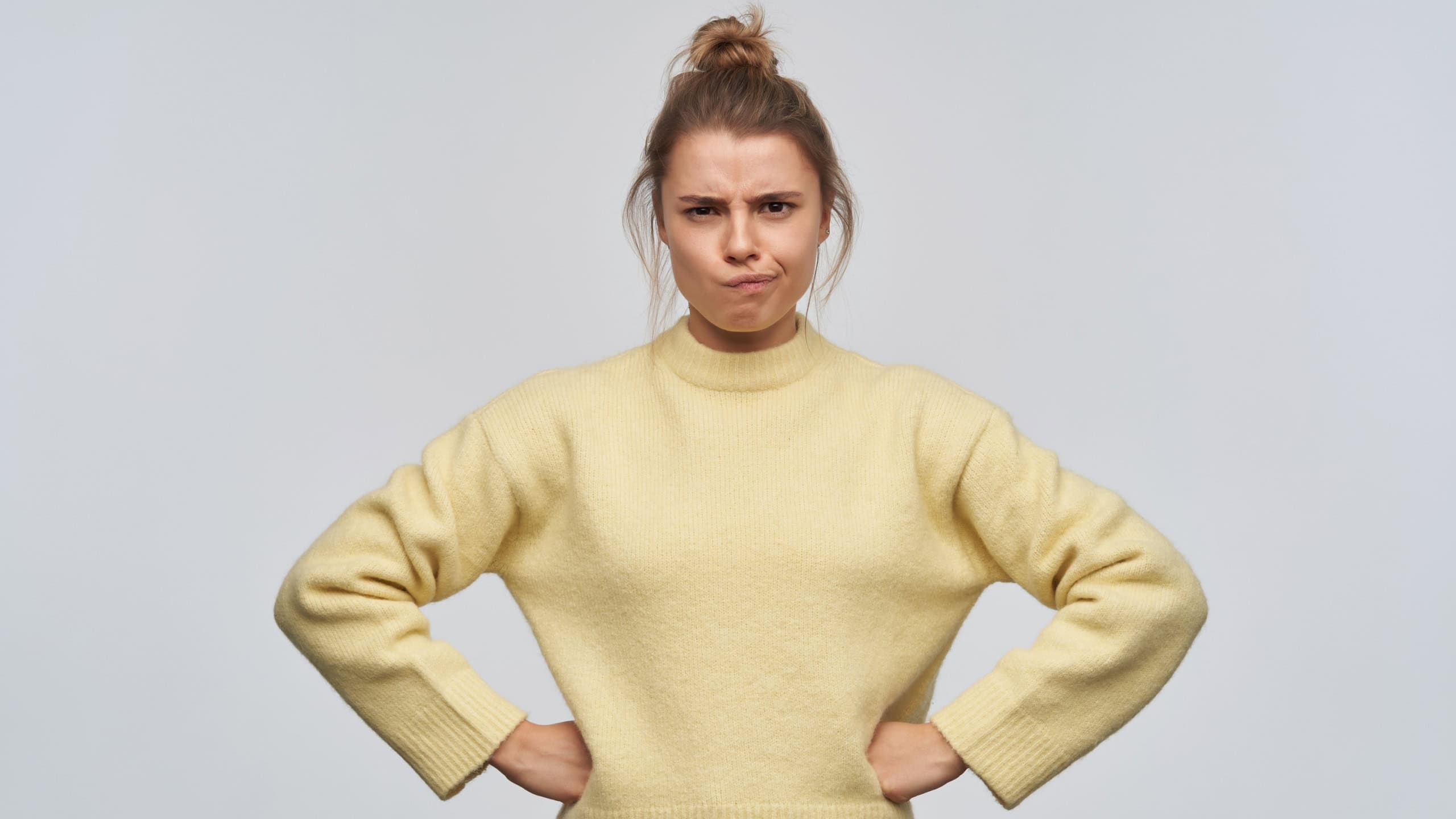 Teenage girl, unhappy looking woman with blond hair gathered in bun. Wearing yellow sweater. Frowns her face and puts her hands on her hips. Watching at the camera, isolated over white background