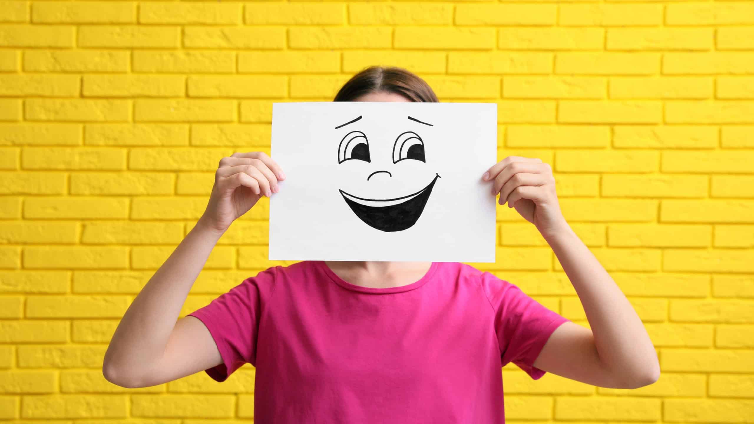 Woman hiding behind sheet of paper with laughing face against yellow brick wall