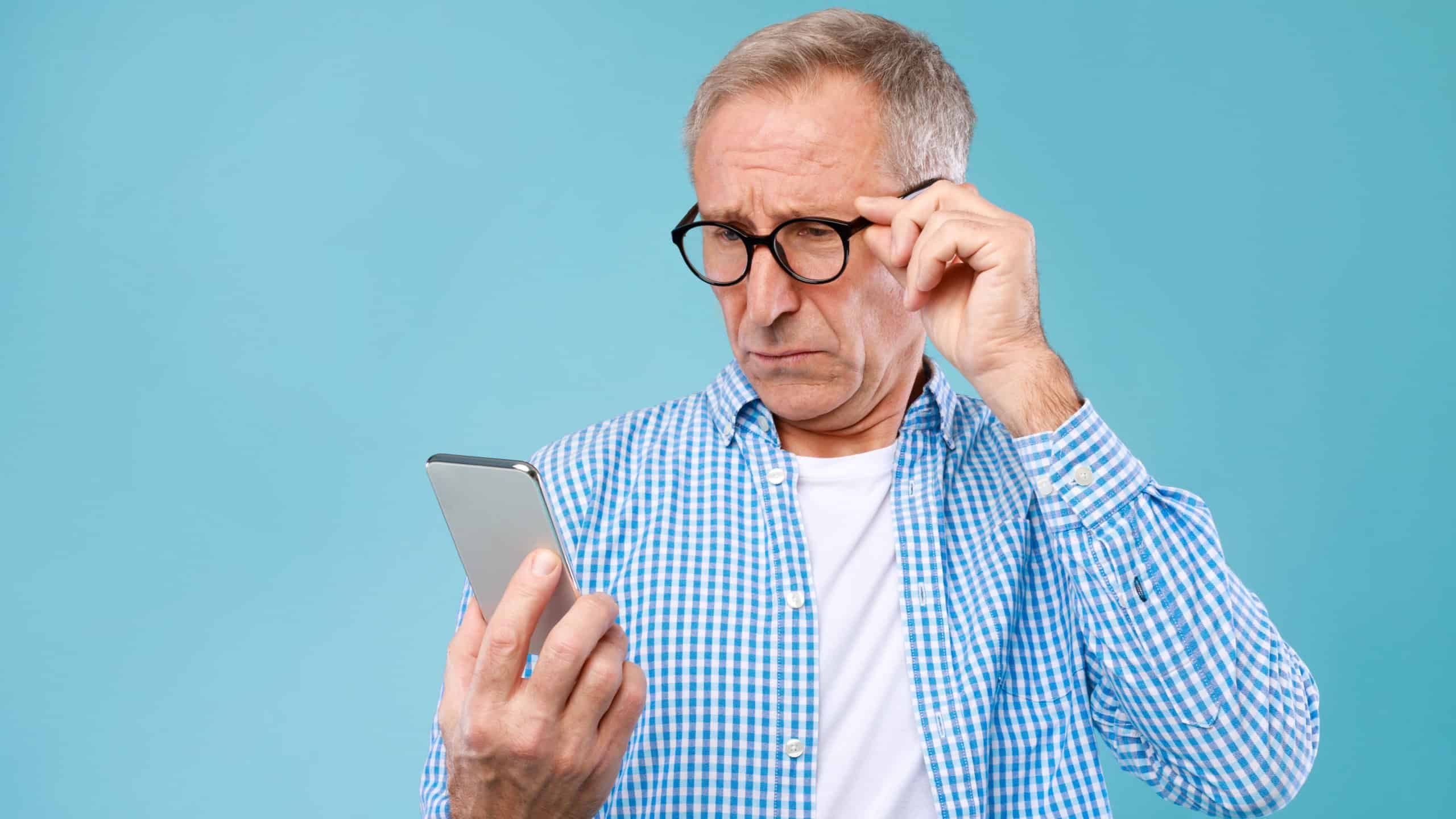 Bad News Concept. Upset Mature Man Holding Smartphone, Looking At Mobile Phone Screen With Worried Expression, Touching Glasses, Sad Adult Male Reading Unplesant Message, Blue Studio Background