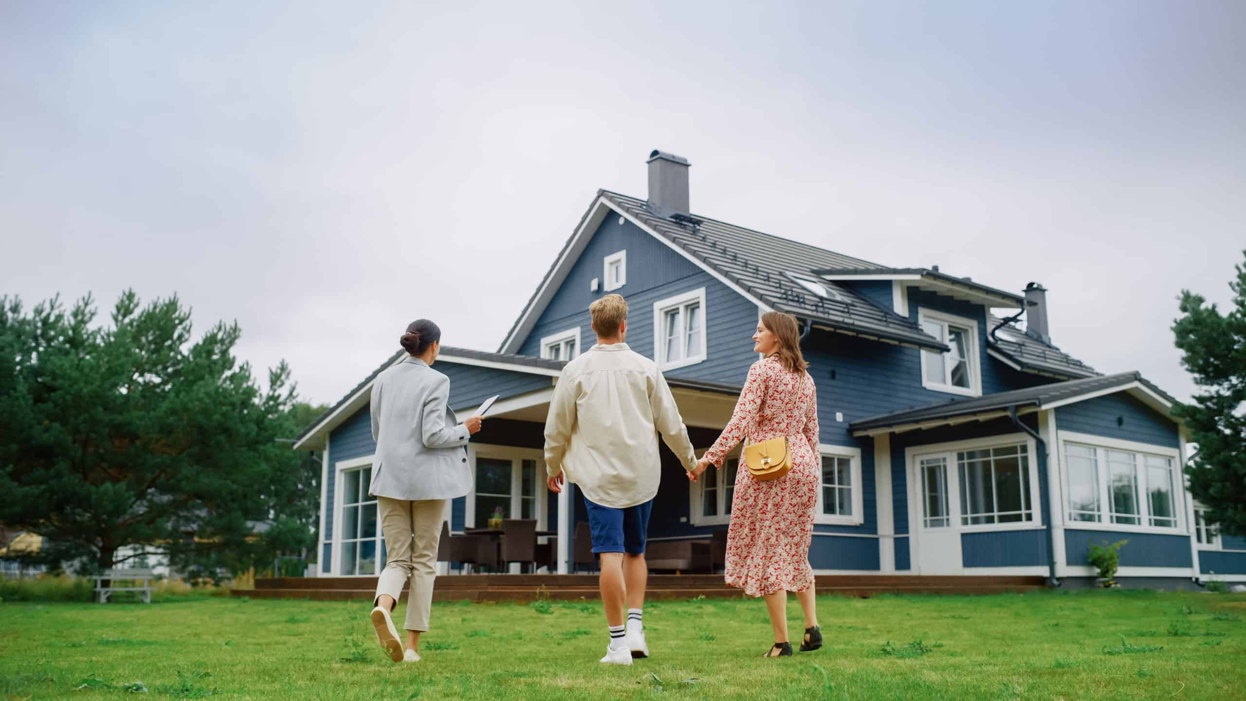 Real Estate Agent Showing a Beautiful Big House to a Young Successful Couple. People Standing Outside on a Warm Day on a Lawn, Talking with Businesswoman, Discussing Buying a New Home.