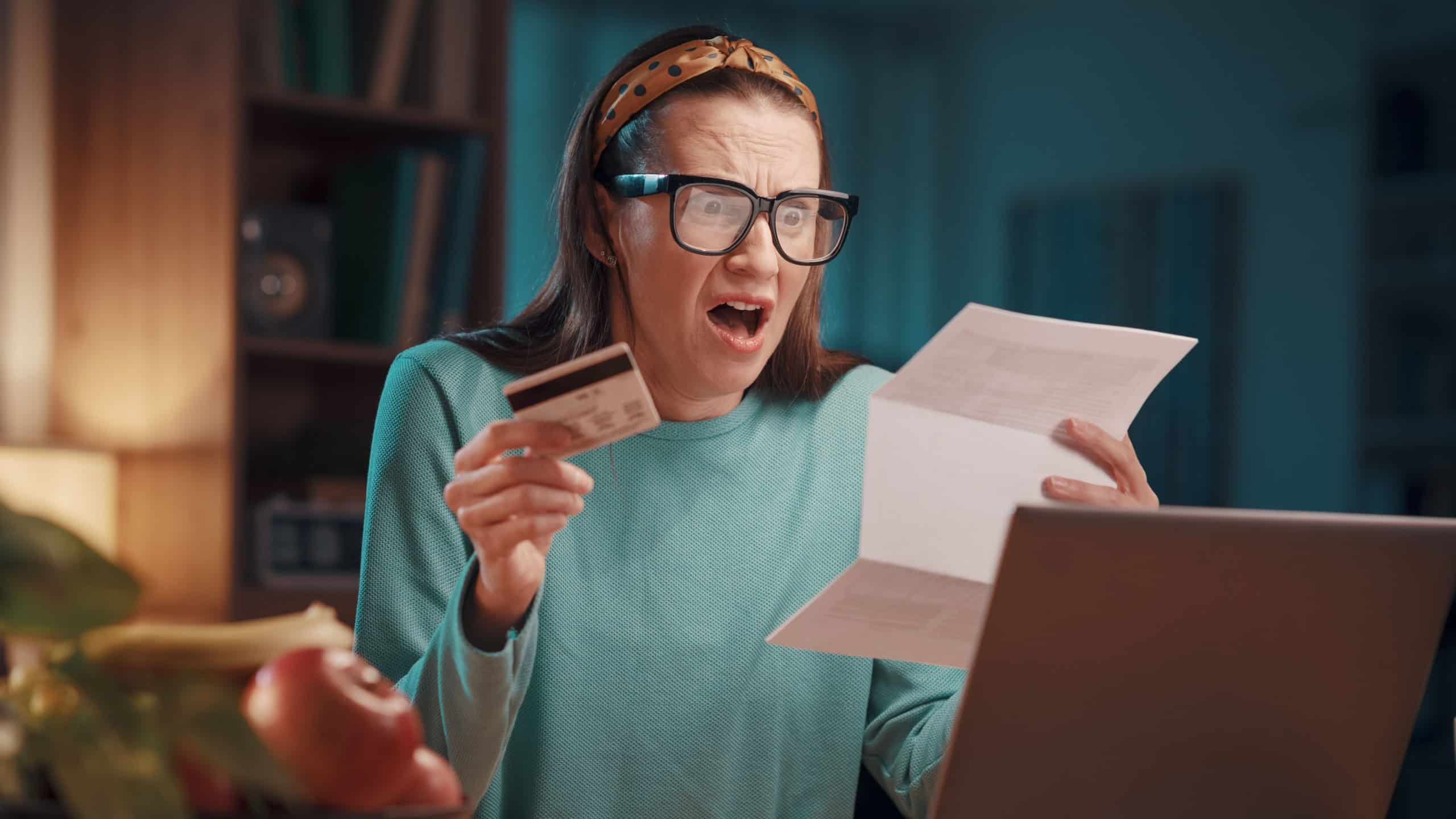 Desperate woman sitting at her desk at home and checking an expensive invoice: payments, taxes and bills concept
