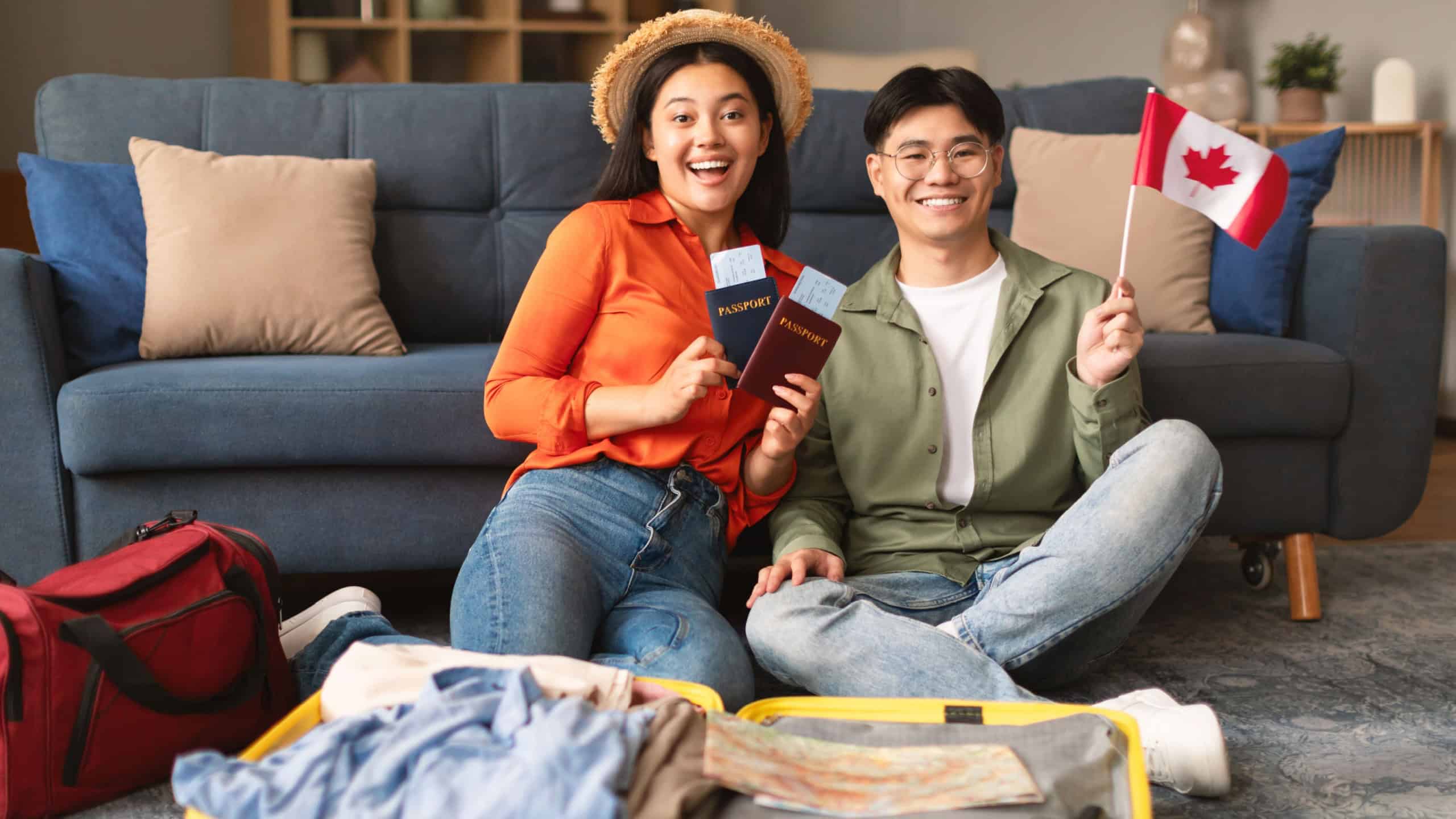 Excited Asian Young Couple Posing Holding Flag Of Canada, Packing Travel Suitcase For New Journey At Home. Spouses Smiling To Camera Preparing For Relocation And Trip Abroad