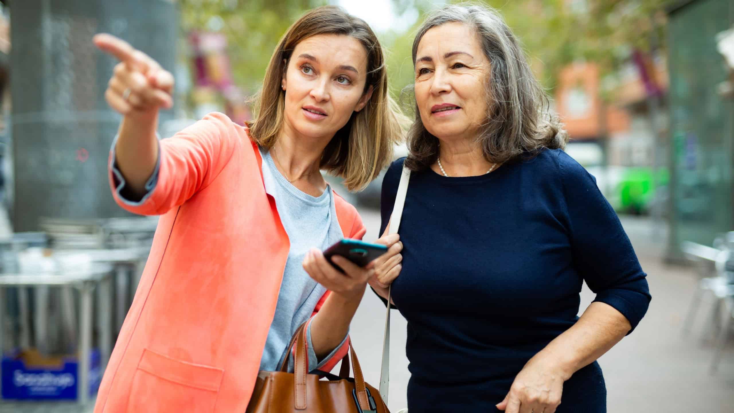 Friendly girl helping elderly woman to find necessary address in city, pointing direction with hand.