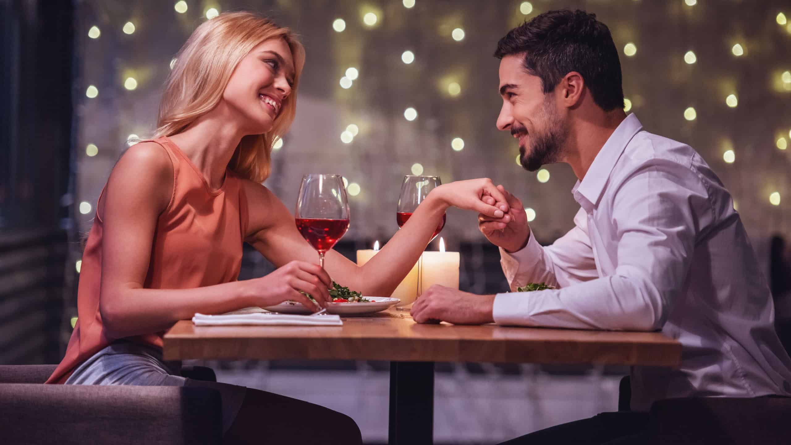 Beautiful young couple is looking at each other and smiling during their date in a restaurant, man is holding his girlfriend's hand