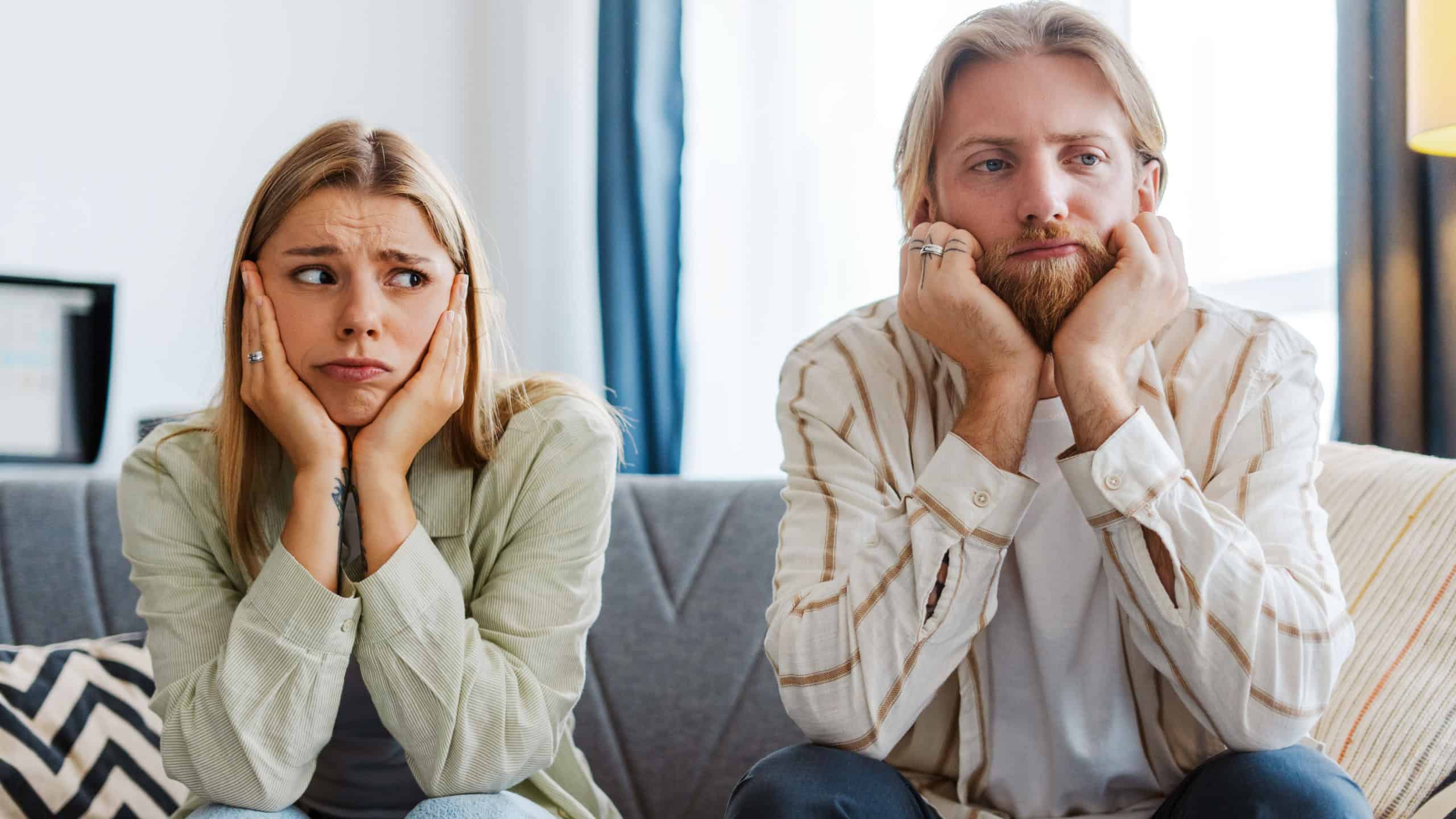 Sad, bored couple sits on a cozy sofa at home, showing signs of relationship struggles through their tired, frustrated body language