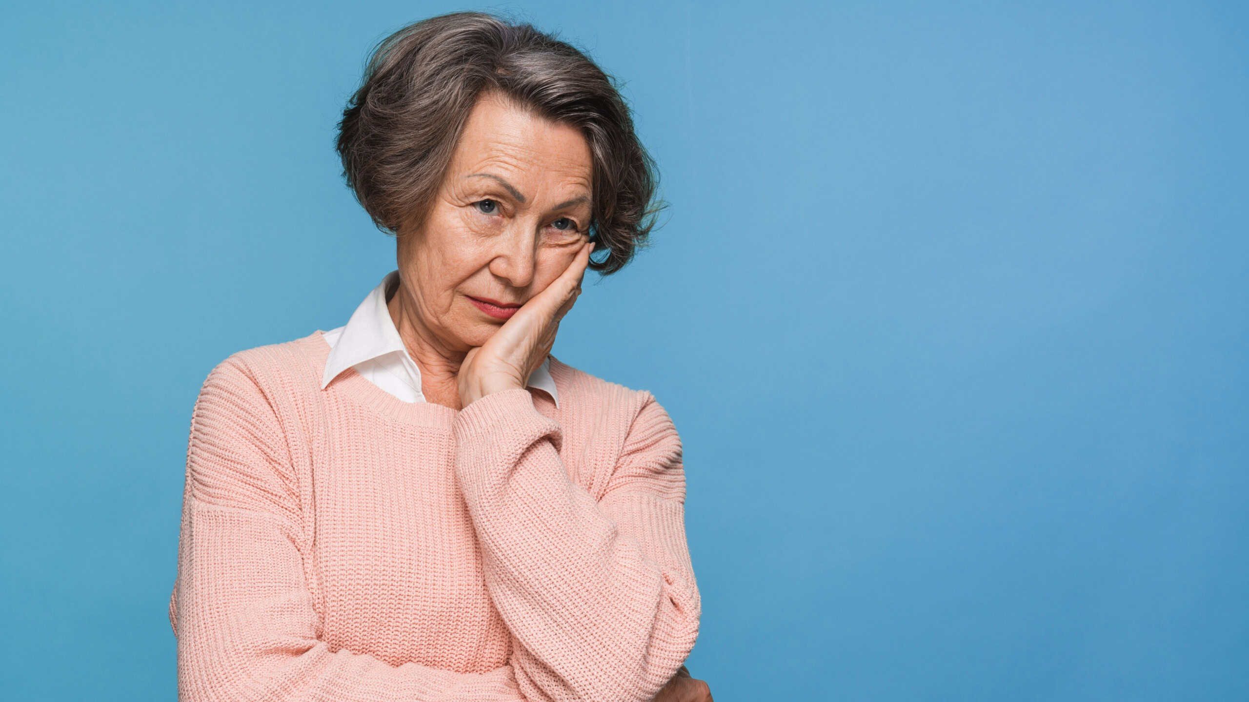 Unhappy sad senior woman suffering from terrible toothache. Frustrated female pensioner in blue shirt holding hand on her face having worried uneasy facial expression, thinking about her future.