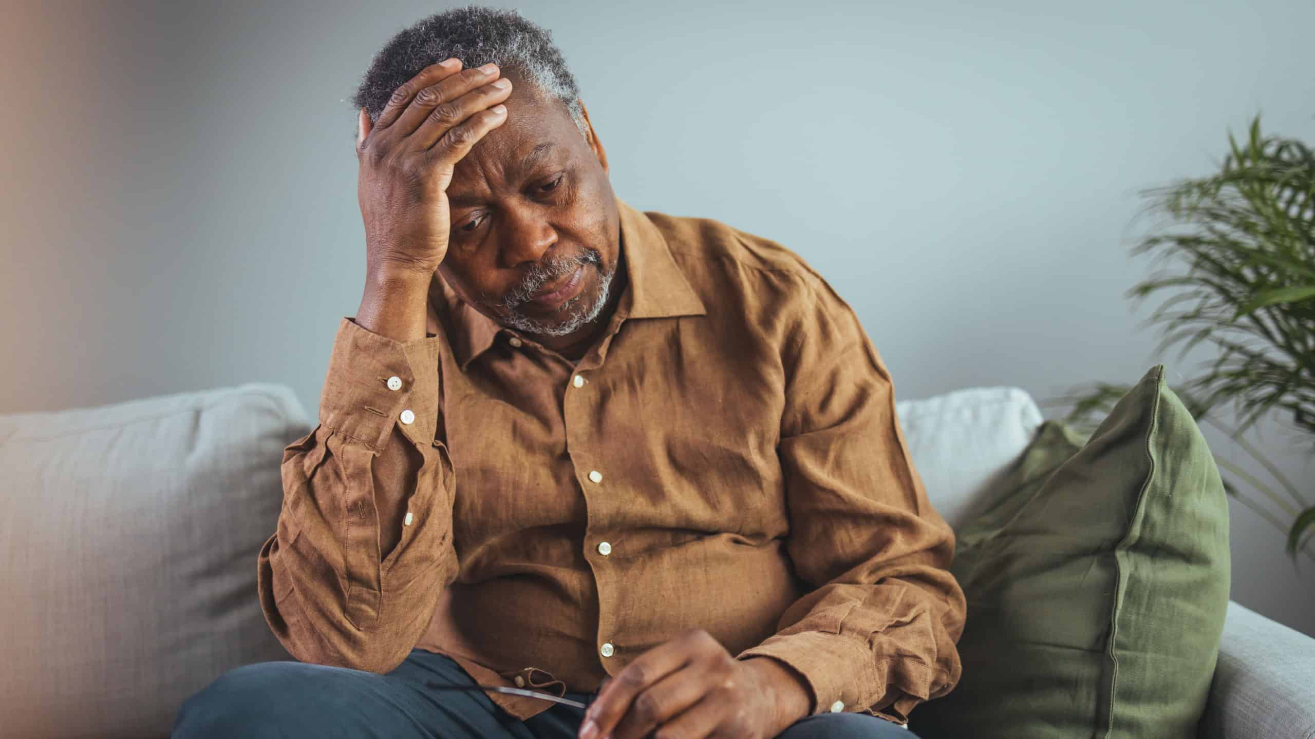 Elderly Black Man Experiencing Discomfort on Couch. Senior black male grips his glasses, showing signs of distress while seated alone on a sofa, reflecting potential health concerns in a home setting.