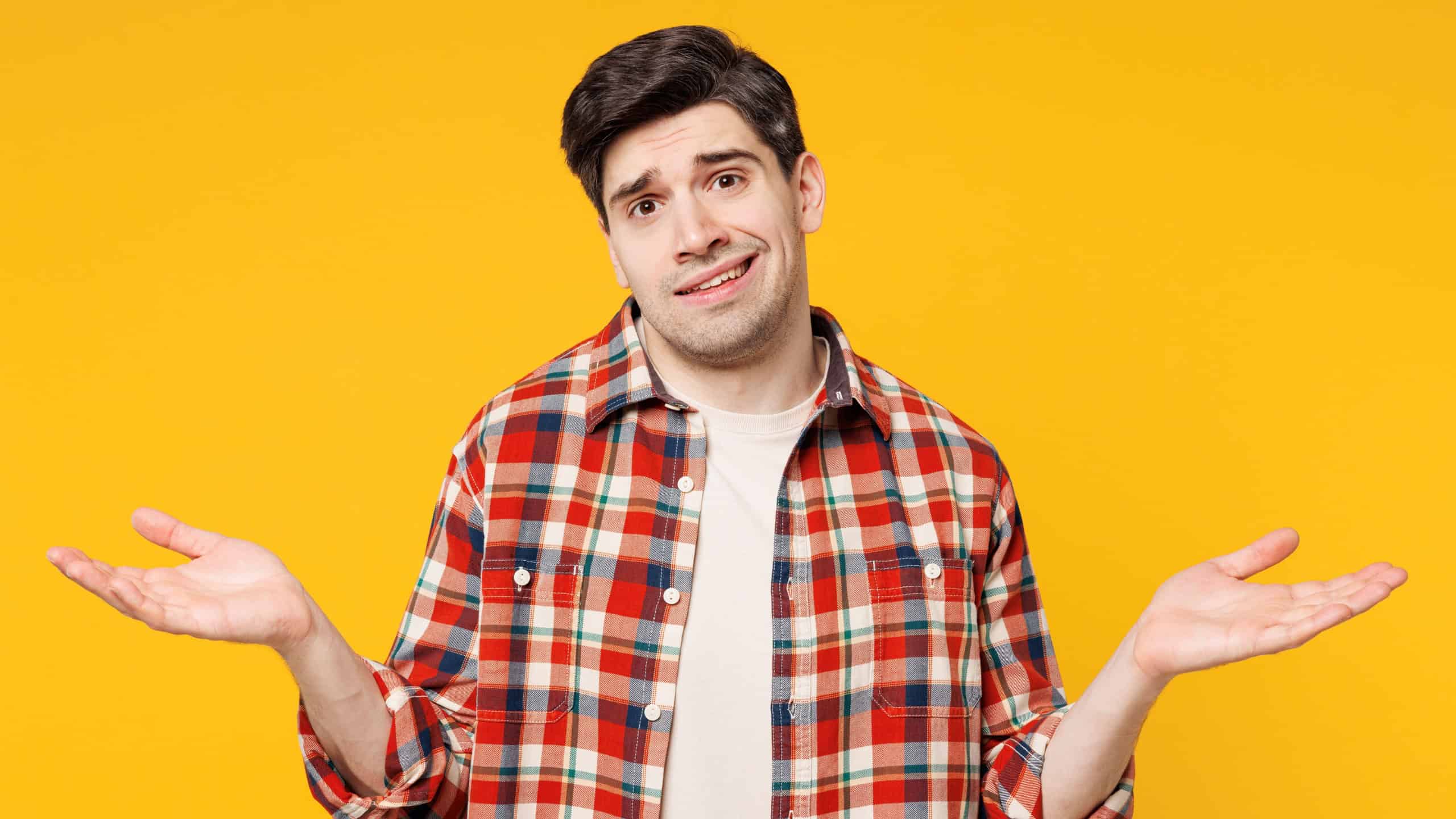 Young sad man he wears red checkered shirt casual clothes shrugging shoulders looking puzzled, have no idea spread hands isolated on plain yellow orange background studio portrait. Lifestyle concept