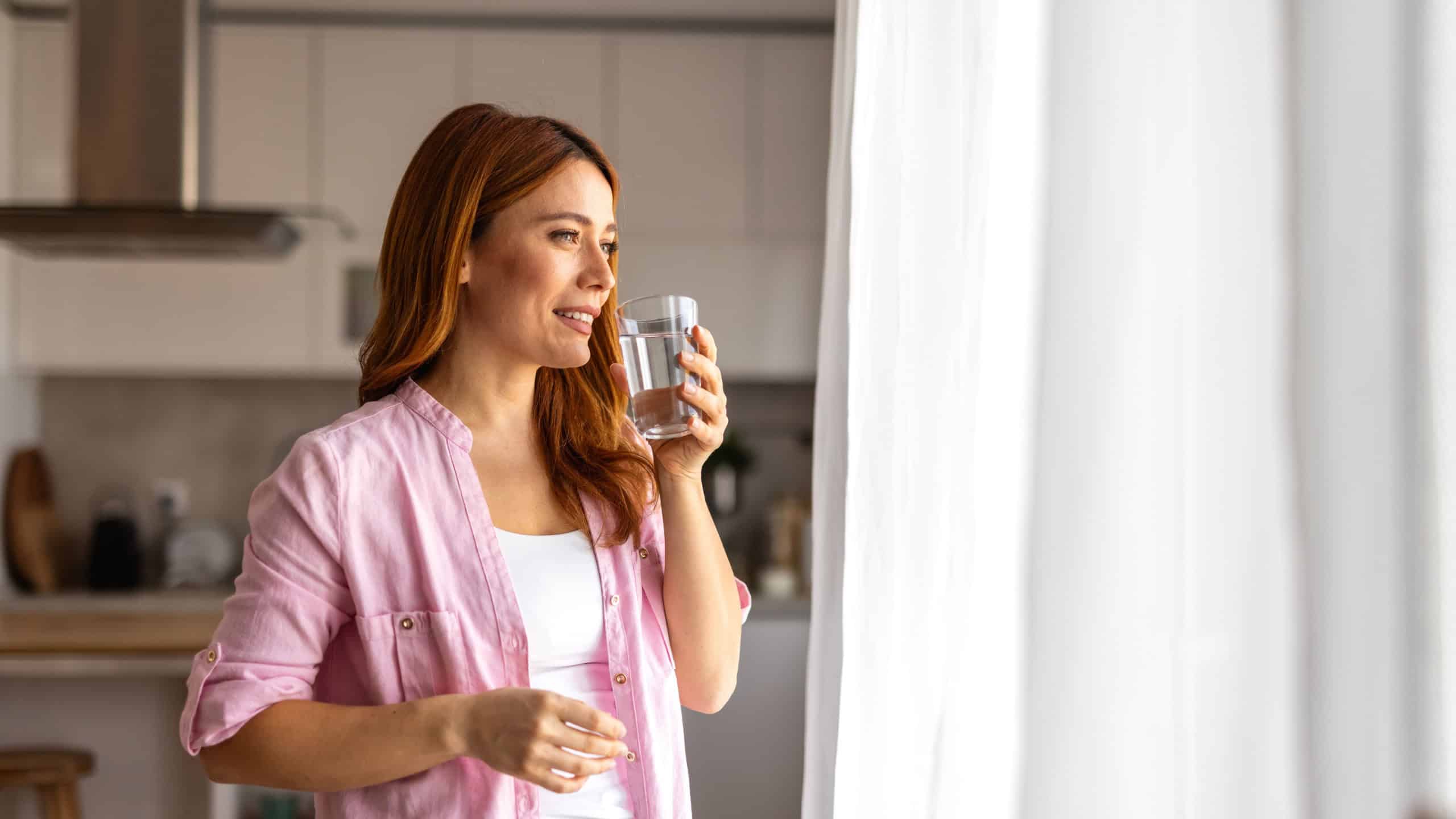 Standing by the window a woman drinks a fresh glass of water, savoring the moment of relaxation. Focused on hydration and well being, she enjoys a quiet break in her bright and inviting home