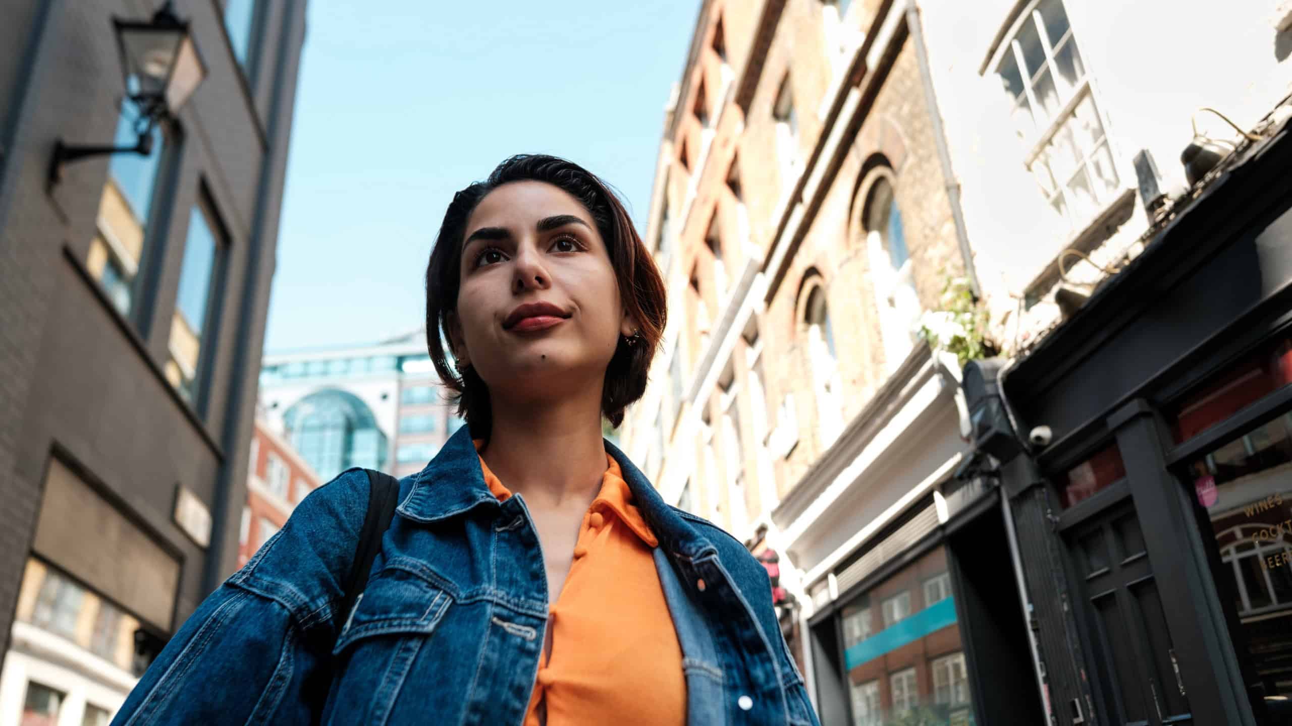 Stylish young woman wearing denim jacket and orange shirt is walking in london city center during a sunny day, enjoying her vacation in europe