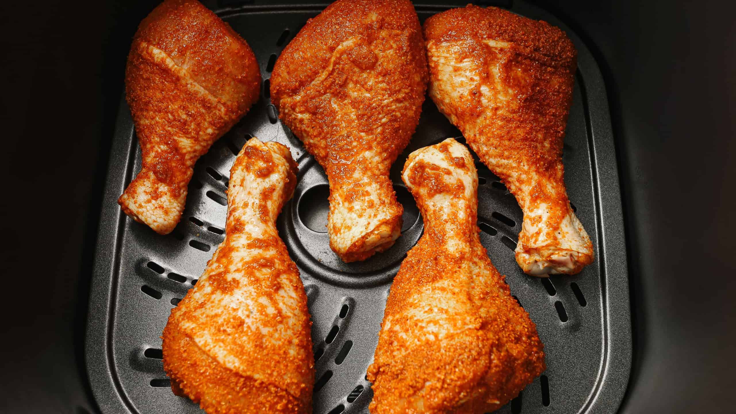 Chicken drumsticks with seasoning in air fryer basket ready for cooking. Spiced poultry meat prepared for roasting. Homemade family meal idea. Small electric oven for quick dinner.