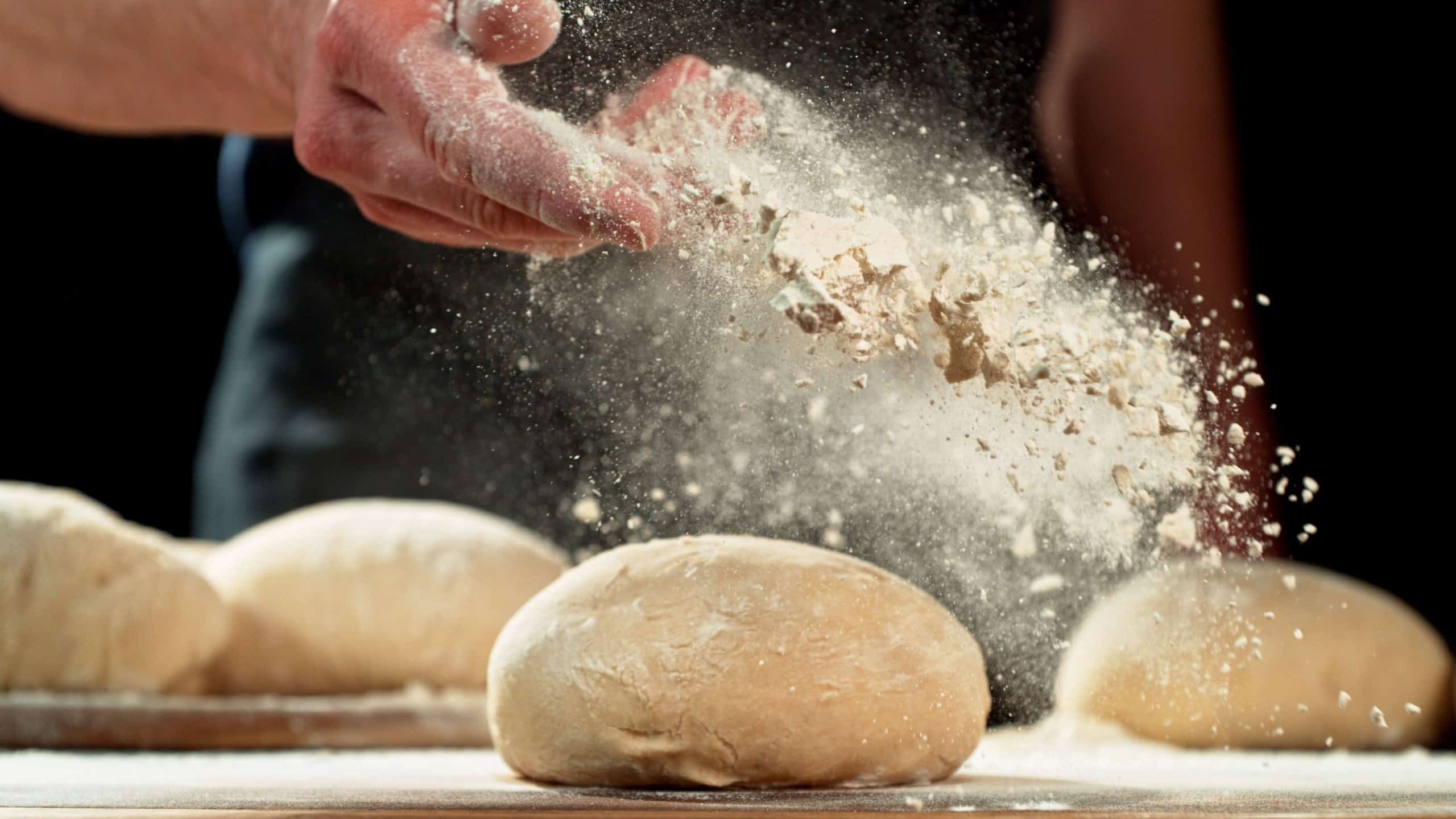 Close up of baker’s hand tossing flour over prepared risen dough on wooden table. Cinematic baking concept, artisan bread preparation and texture detail.