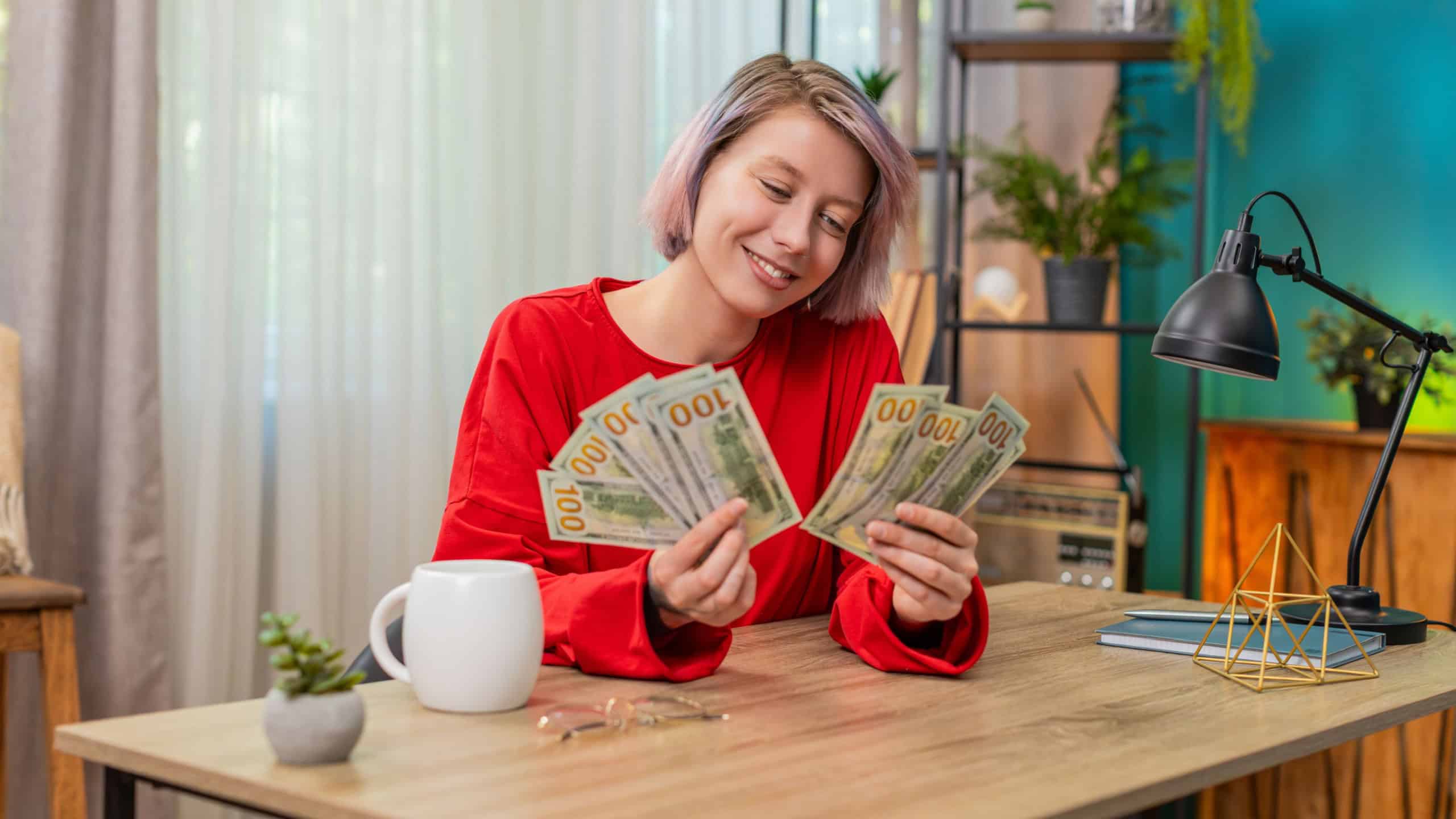 Young woman counting dollars cash money at home table, widely smiling nodding satisfied, expressing wealth. Caucasian girl cheerfully demonstrating joyful financial success and prosperity achievement.
