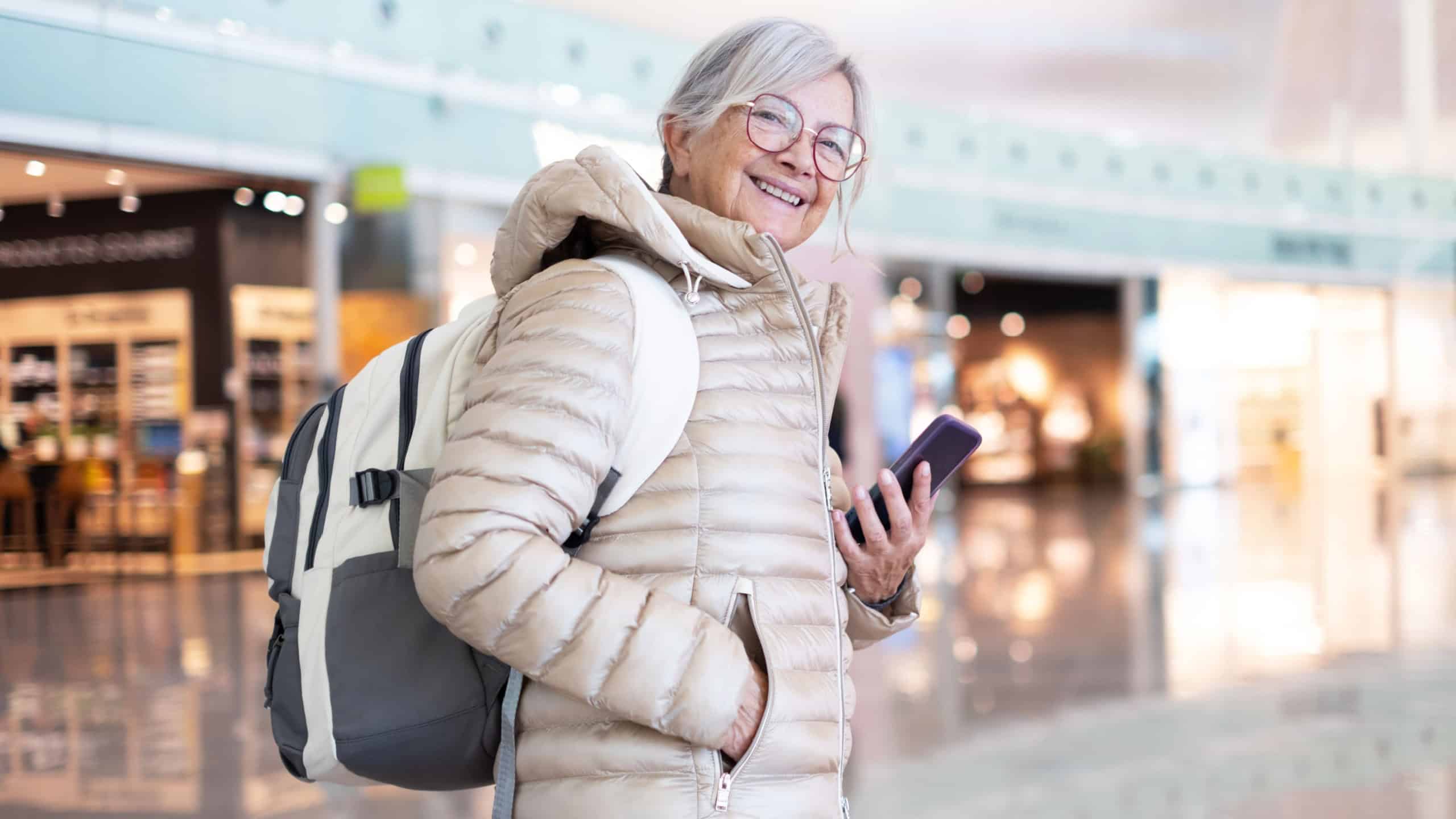 Confident senior woman traveler wearing a warm jacket and backpack, smiling while checking her smartphone in a bright modern airport terminal, conveying independence, travel readiness and joyful anticipation.