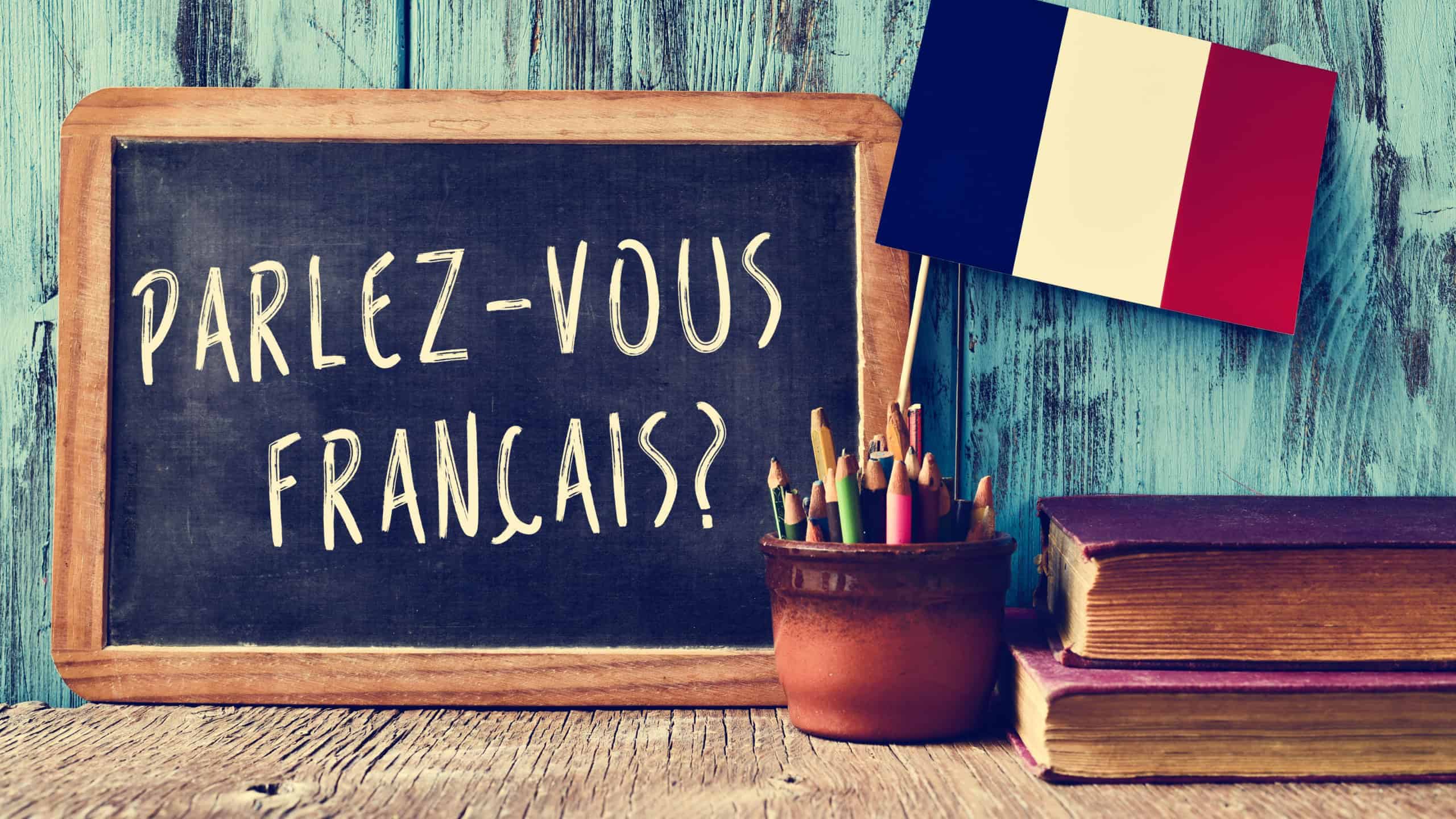 a chalkboard with the question parlez-vous francais? do you speak french? written in french, a pot with pencils and the flag of France, on a wooden desk