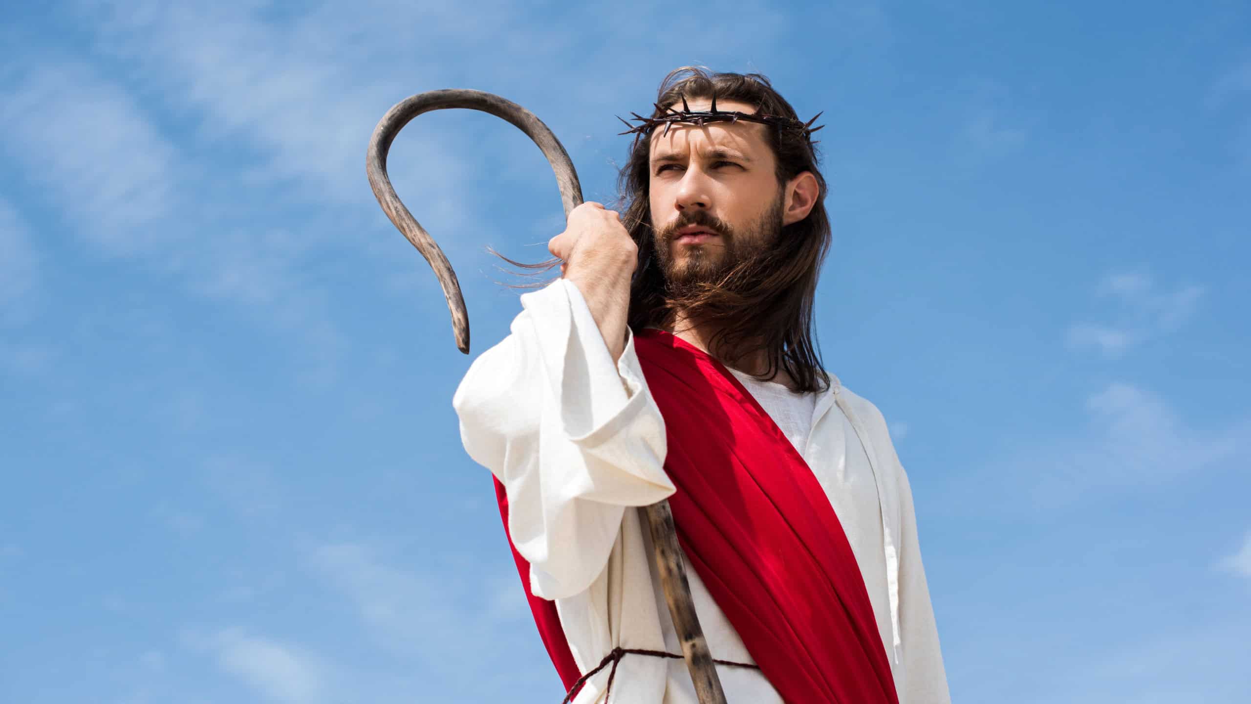 Jesus in robe, red sash and crown of thorns standing with wooden staff against blue sky and looking away