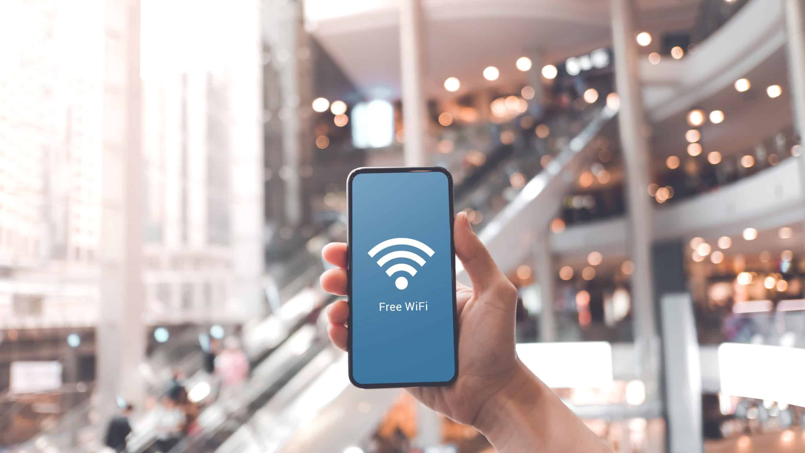 Hand holding smartphone with free wifi icon on blurred shopping mall background.